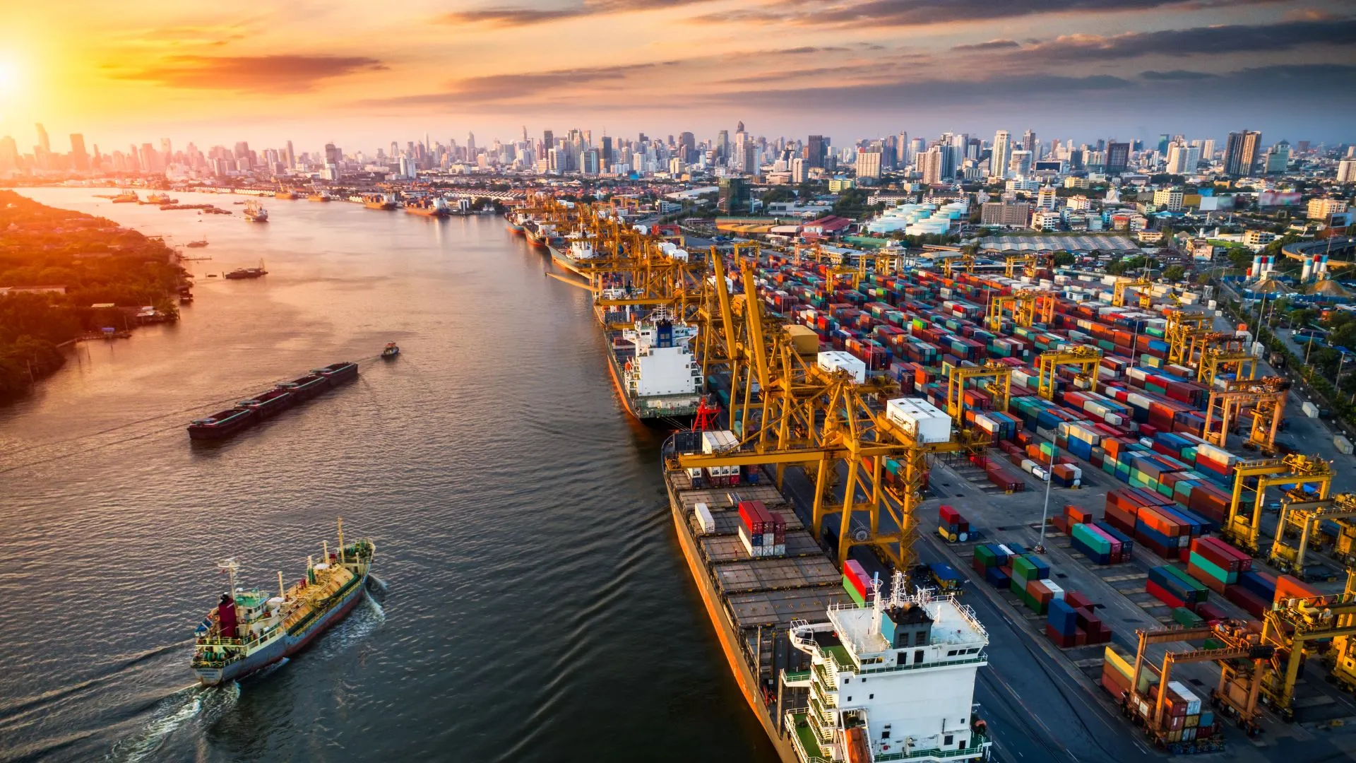 Aerial view of a busy shipping port with container ships docked, colorful shipping containers, cranes, and a city skyline at sunset.