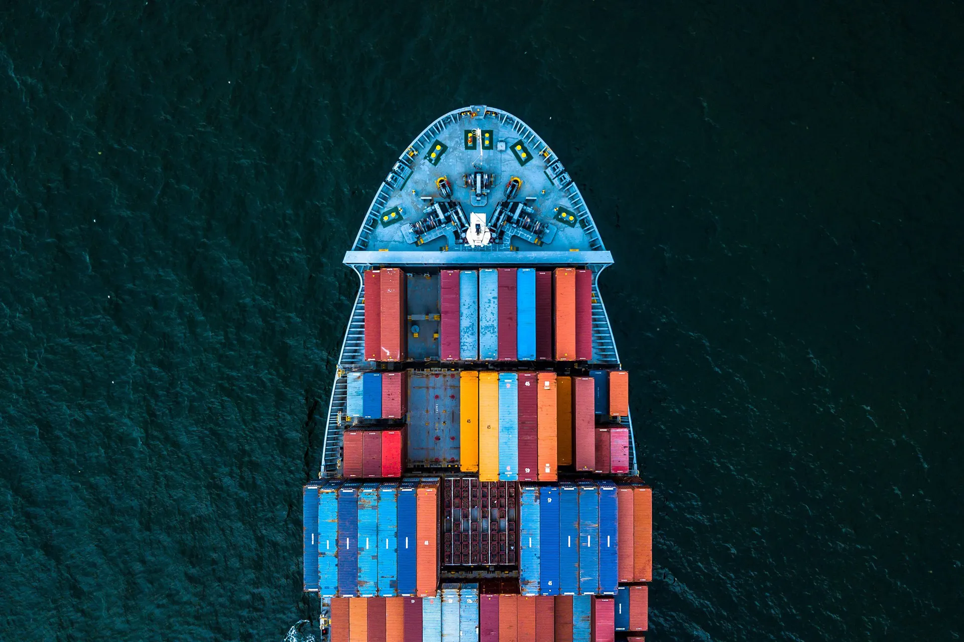 Aerial view of a large container ship loaded with colorful shipping containers sailing on dark blue water.