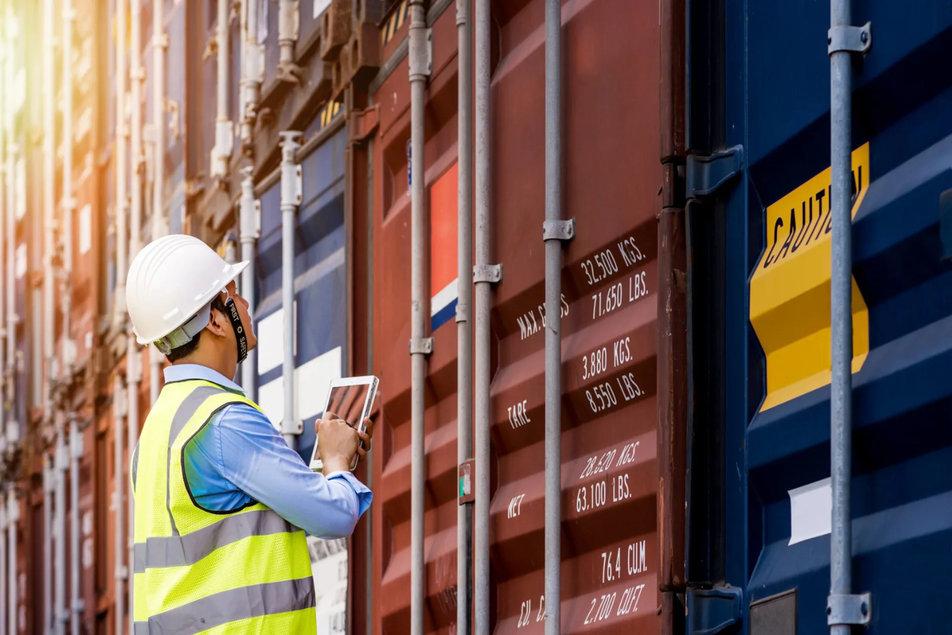 Worker in safety vest and helmet inspecting shipping containers with a tablet in hand.
