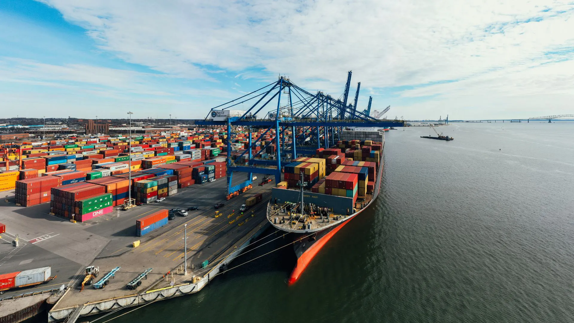 A large container ship docked at a busy port terminal with numerous colorful shipping containers and blue cranes.