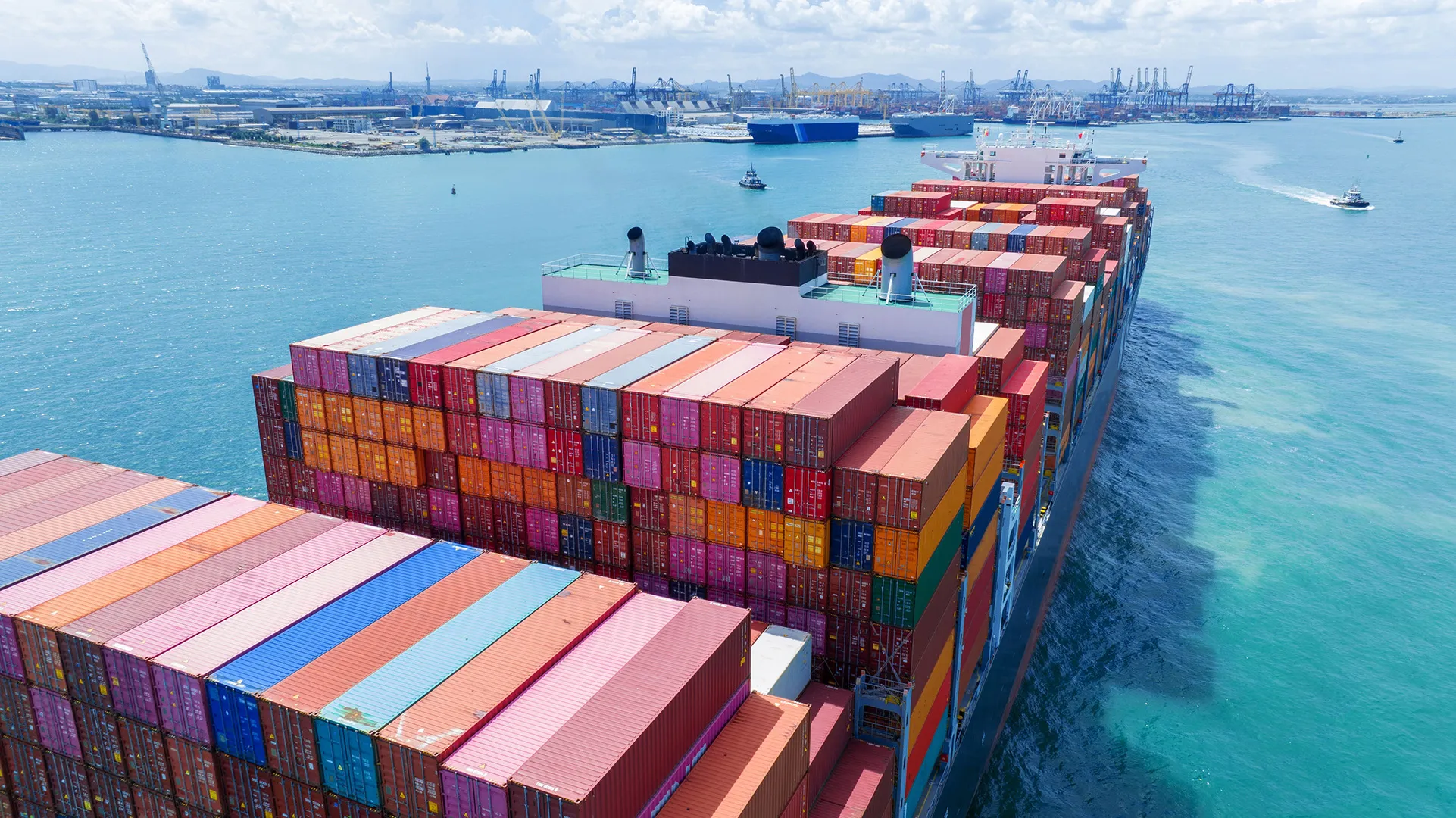 A large cargo ship loaded with colorful shipping containers sailing in a harbor with cranes and other ships in the background.