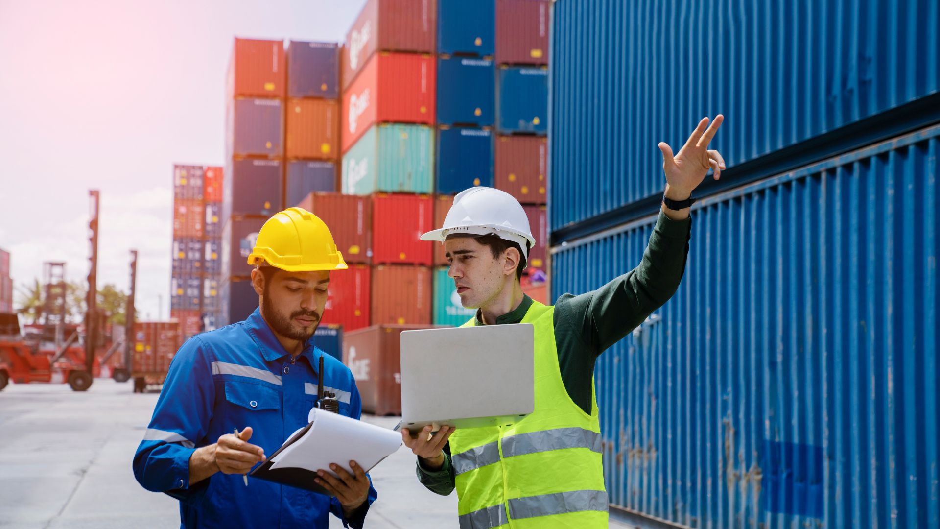 Two workers in safety gear inspecting documents and pointing at shipping containers in a port yard.