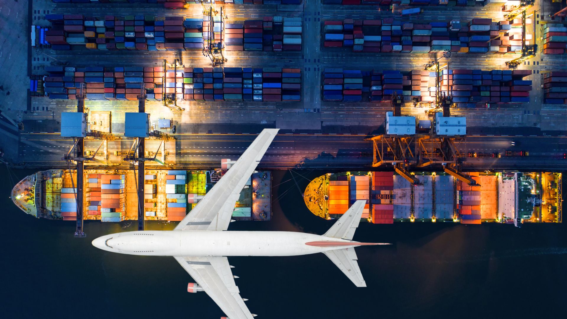 Aerial night view of a large white airplane flying over an illuminated cargo port with container ships and stacked shipping containers.