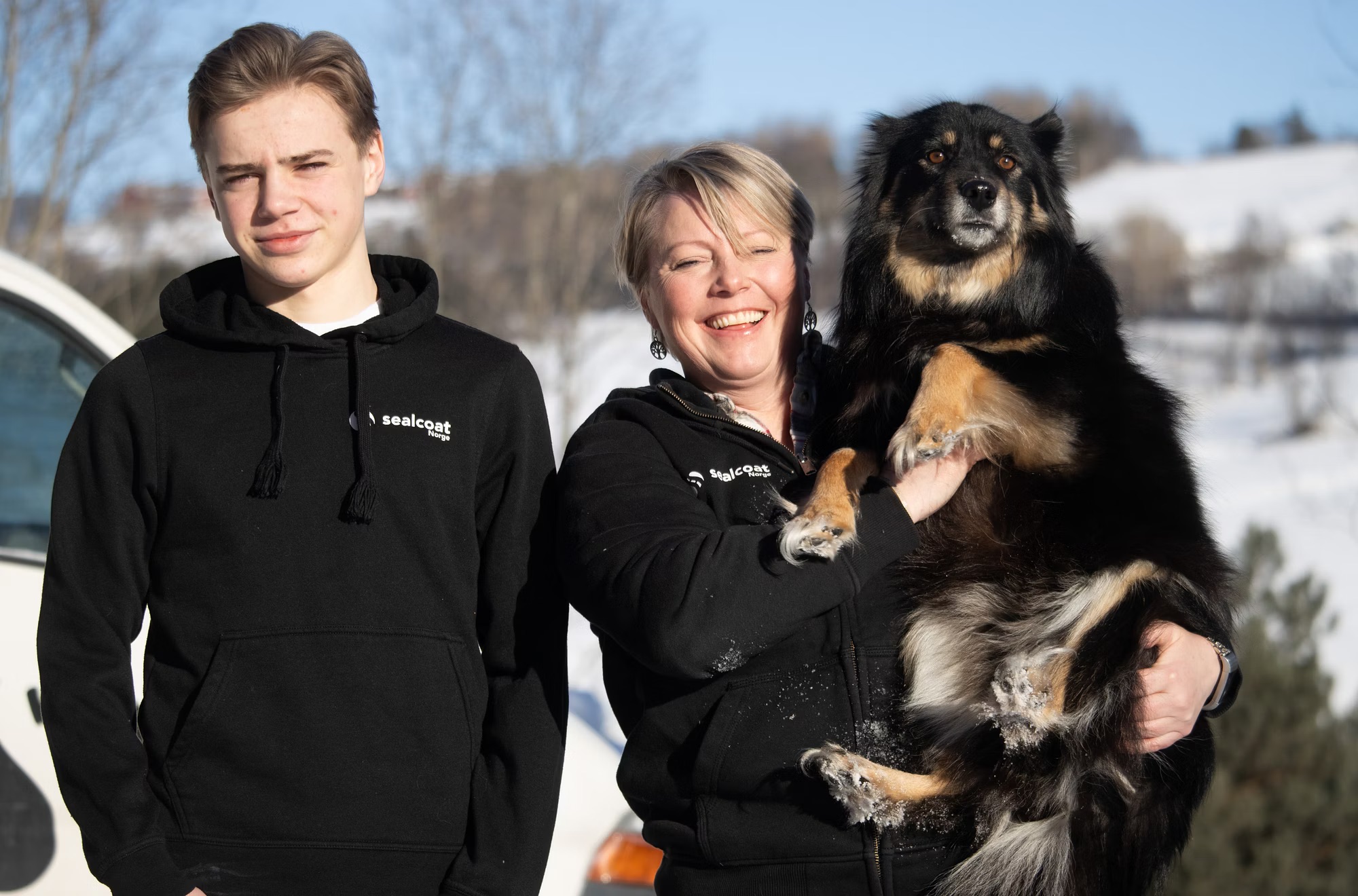 To personer i svarte sealcoat klær, kvinnen holder en stor svart og brun hund utendørs i vinter.