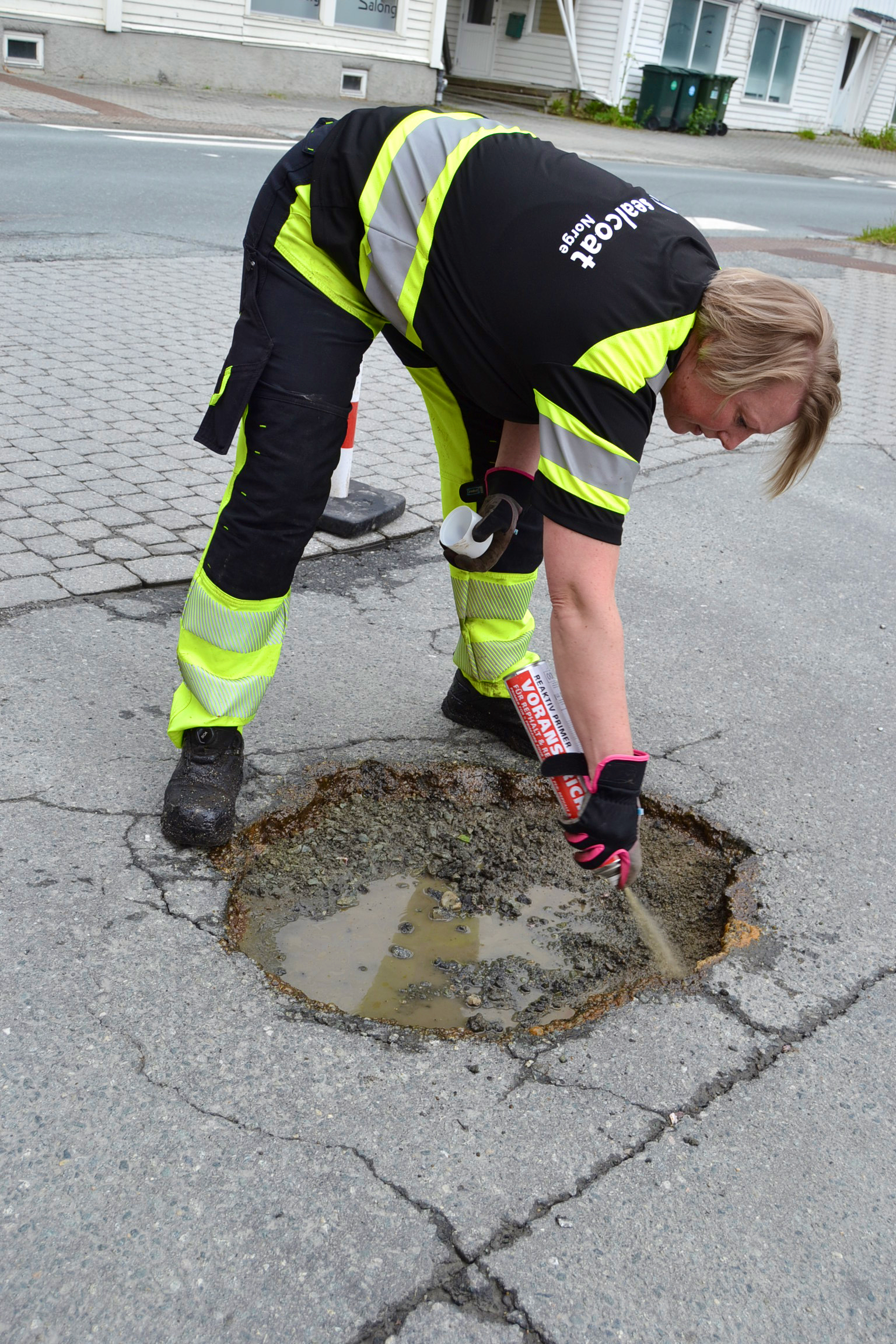 Arbeider i gul og svart vernedrakt reparerer en stor vannfylt hull i asfaltveien.