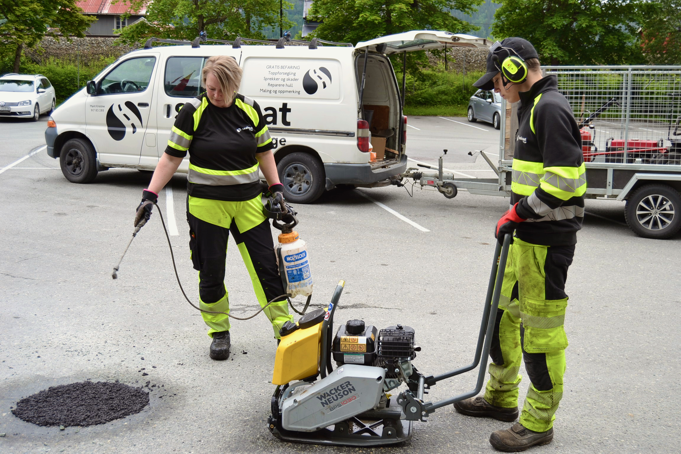 To arbeidere i verneutstyr fyller asfalt i en parkeringsplass med asfaltmaskin og sprøyteflaske.