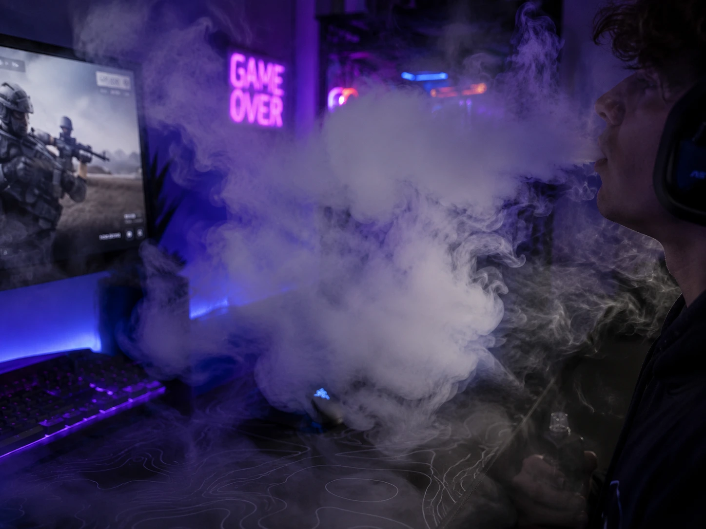Gamer wearing a headset exhaling a large cloud of vape smoke while playing a first-person shooter game at a neon-lit PC gaming setup.