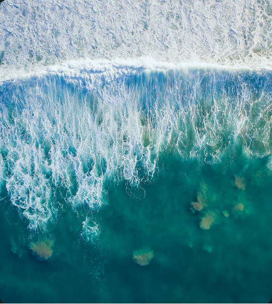 Aerial view of ocean waves breaking and creating white foam above the deep blue and green water with visible underwater rocks.