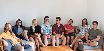 Seven casually dressed people sitting on sofas and a chair against a white wall in a relaxed group setting, with a wooden coffee table in front of them.
