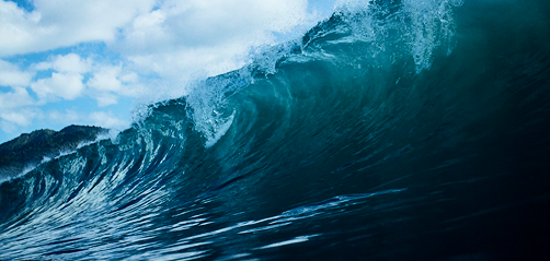 Large ocean wave curling and crashing under a partly cloudy sky with dark blue water.
