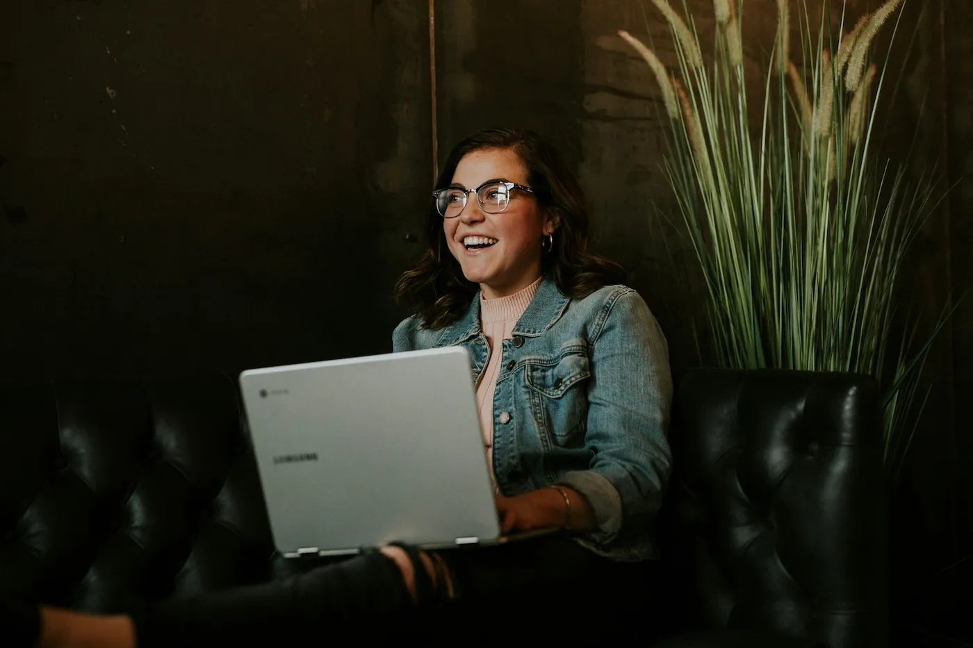 Smiling young woman in glasses and a denim jacket working on a laptop while sitting on a leather sofa, showcasing a relaxed and productive work environment.