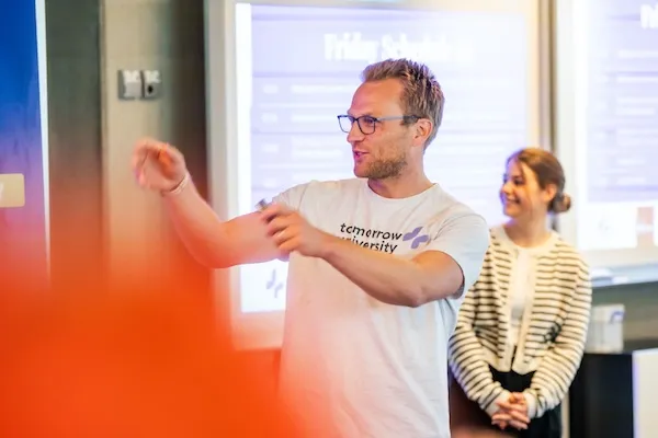 Thomas Funke in a Tomorrow University t-shirt enthusiastically presenting during a community meeting, with a colleague smiling in the background in a modern educational setting.