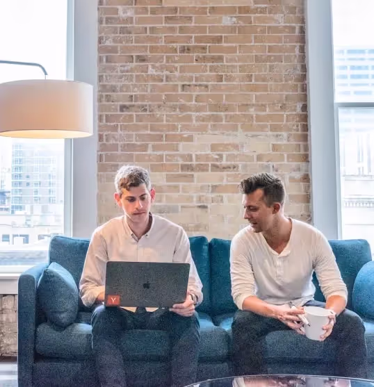Two men sitting on a blue couch in a modern room, one using a laptop and the other holding a white mug.
