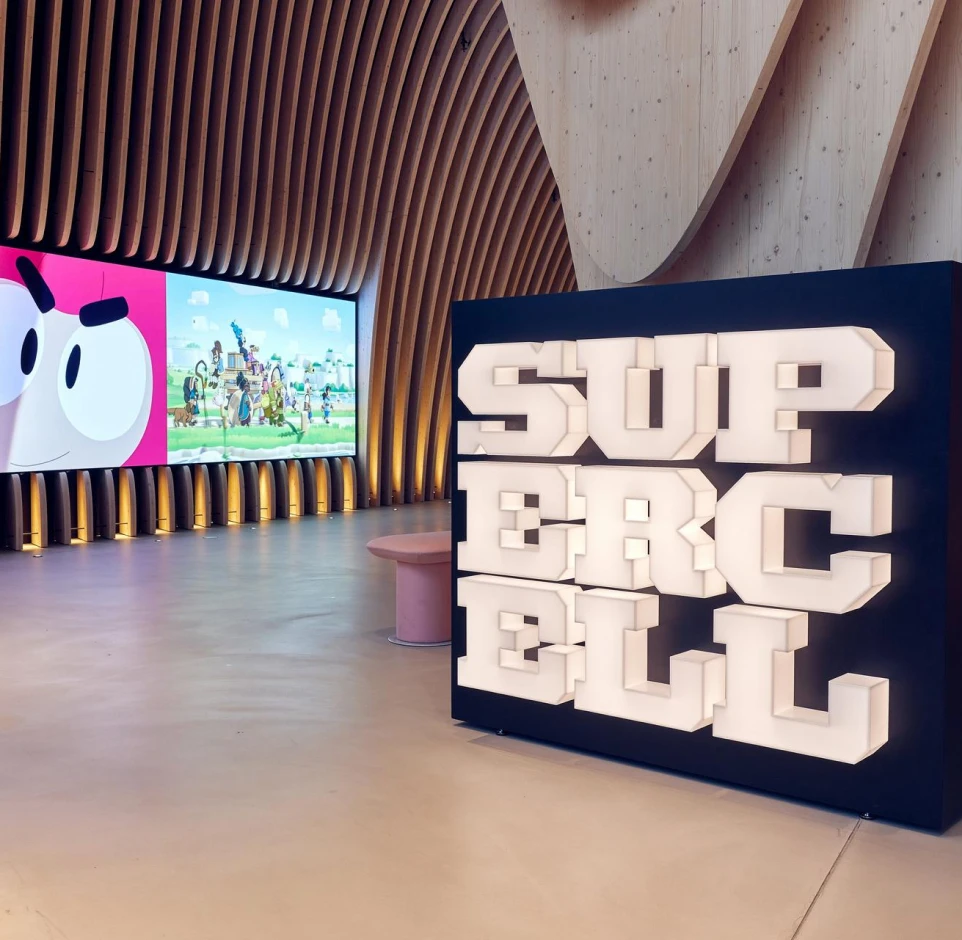 Interior of a modern lobby with wooden curved wall panels, digital screens showing animated characters, and a large illuminated sign reading SUPER CELL.