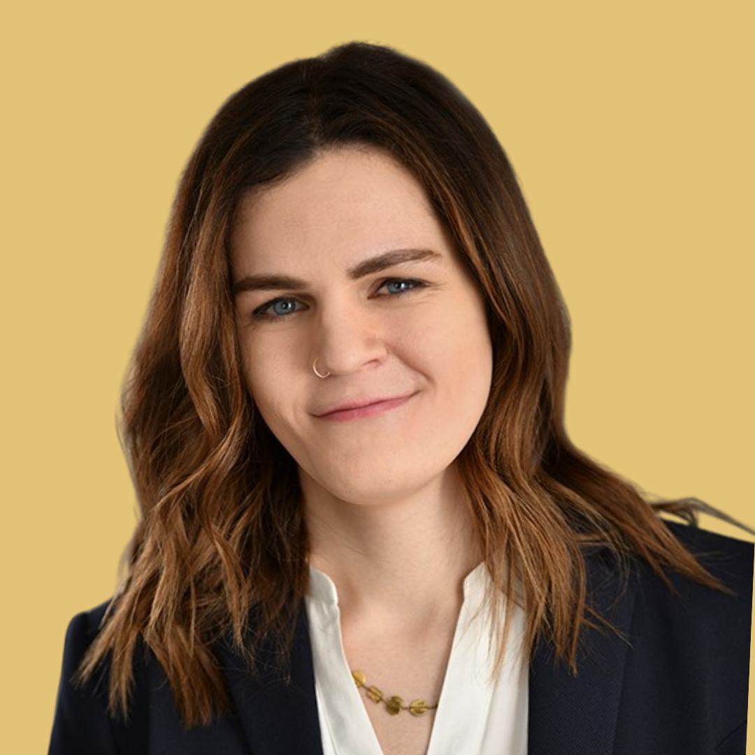 Smiling woman with wavy brown hair wearing a navy blazer, white blouse, and gold necklace on yellow background.