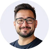 Smiling man with dark hair and beard wearing black rectangular glasses and a dark shirt on a light background.