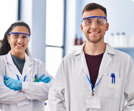 Two smiling scientists wearing white lab coats, safety goggles, and blue gloves standing in a lab with scientific equipment in the background.
