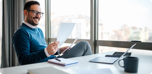 Smiling man with glasses sitting in a modern office near large windows, holding papers, with a laptop, notebook, and coffee mug on the desk.