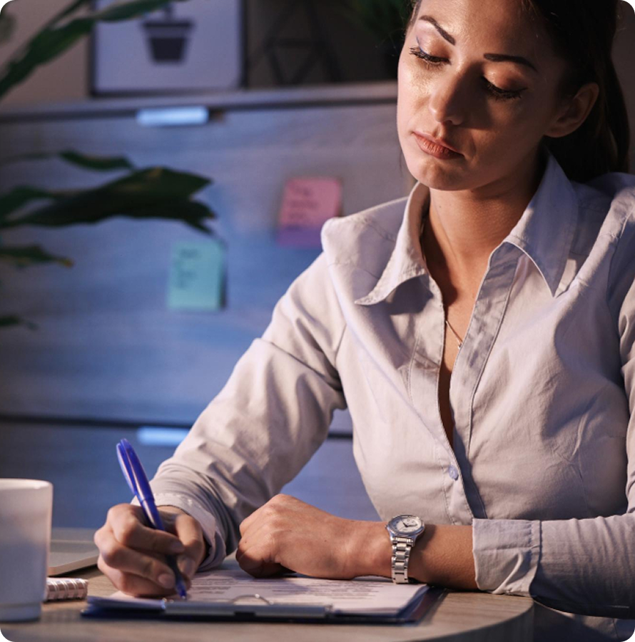 Woman with a light-colored button-up shirt writing with a blue pen on a clipboard at a round table, with sticky notes and a plant in the softly lit background.