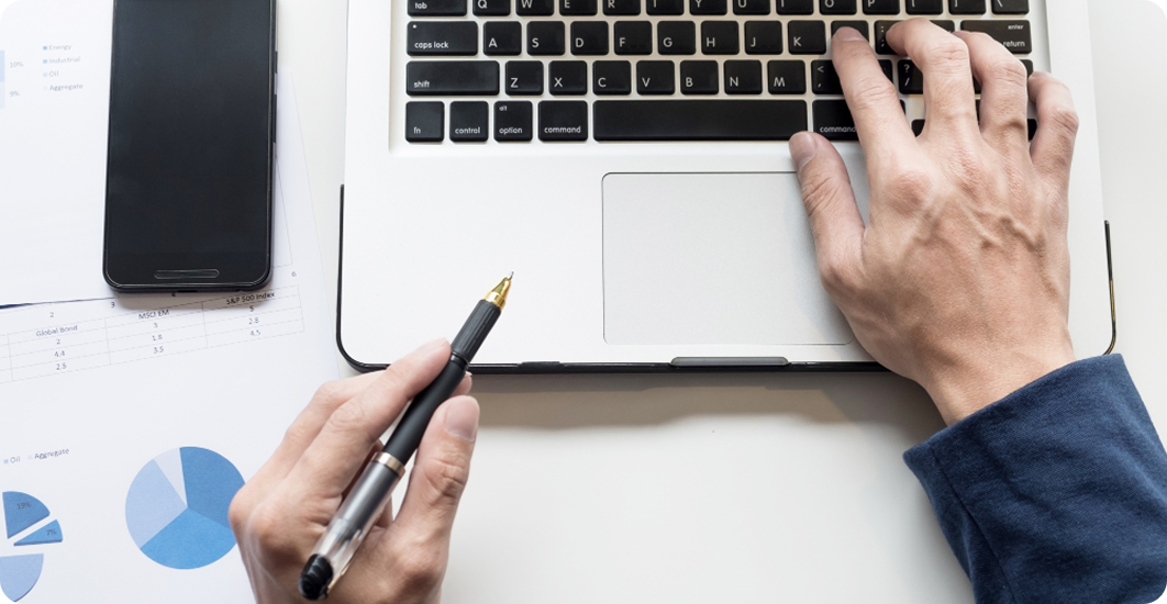 Person's hands using a laptop keyboard with one hand typing and the other holding a black pen above financial documents and a smartphone on a white desk.