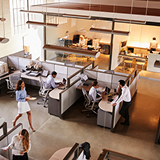 Modern office with employees working in pods and at desks, featuring large windows and overhead pendant lighting.