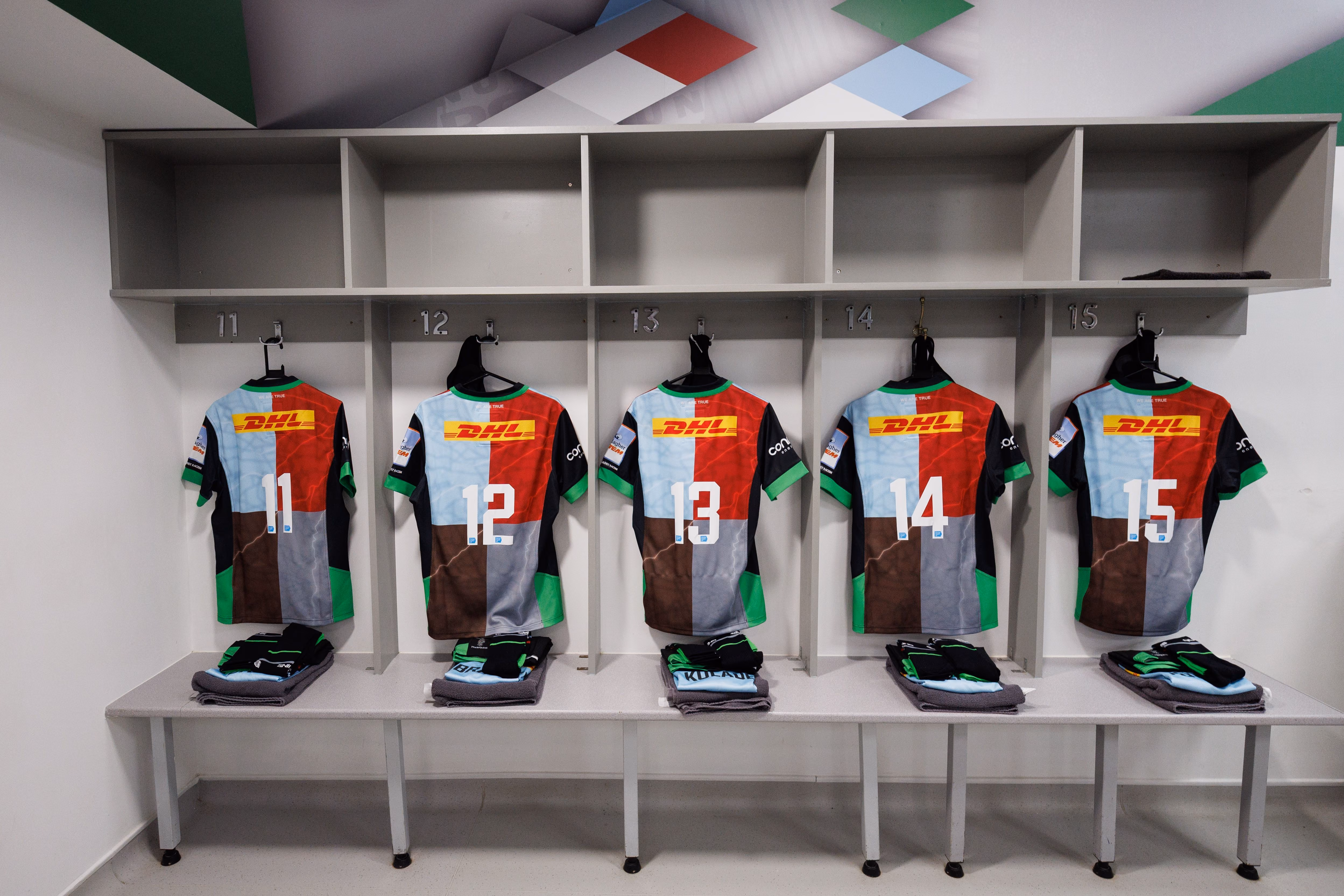 Five multicolored sports jerseys numbered 11 to 15 hanging in a locker room with matching folded uniforms below each jersey.