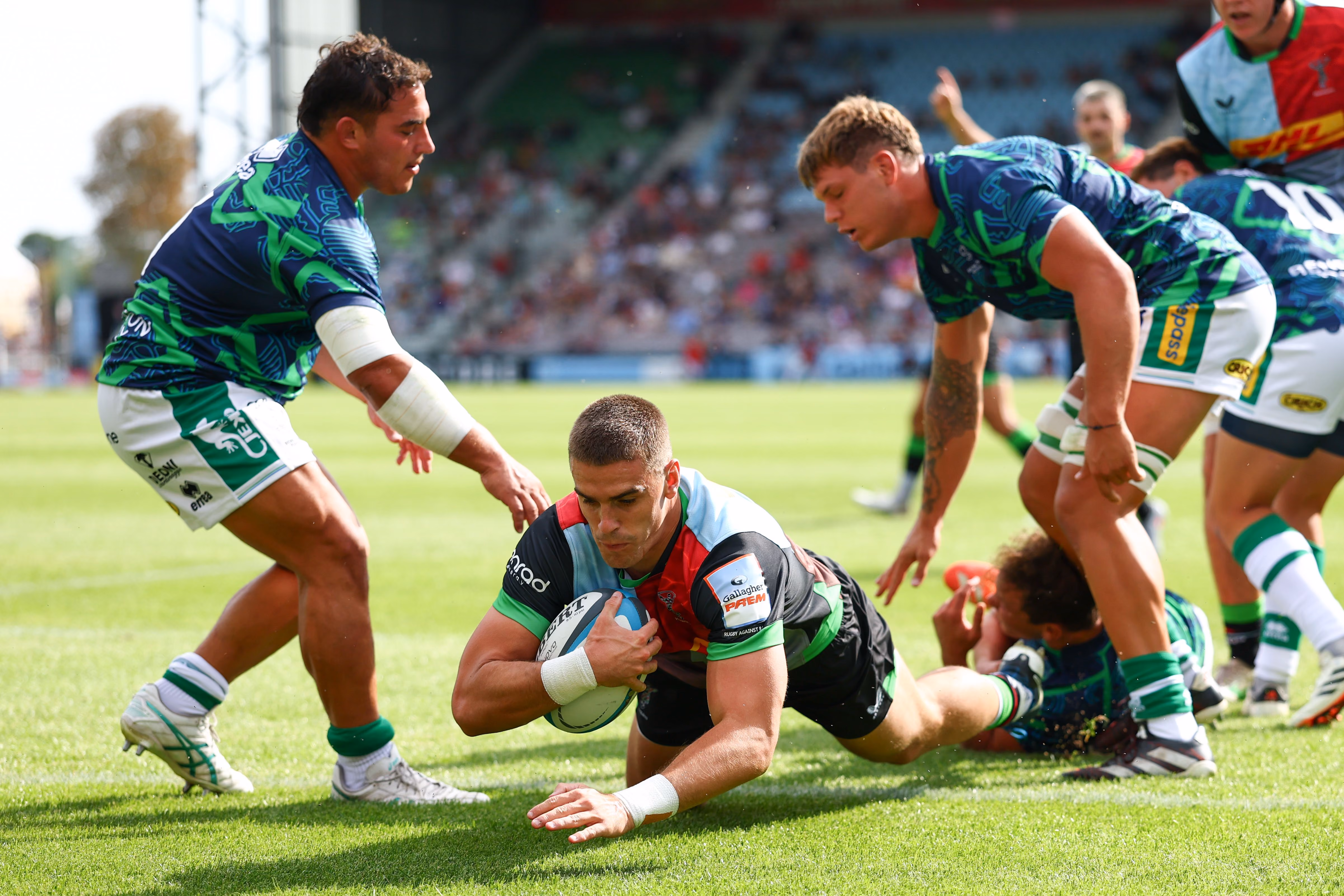 Rugby player in black, red, and light blue jersey diving to score a try while two defenders in blue and green jerseys attempt to stop him.