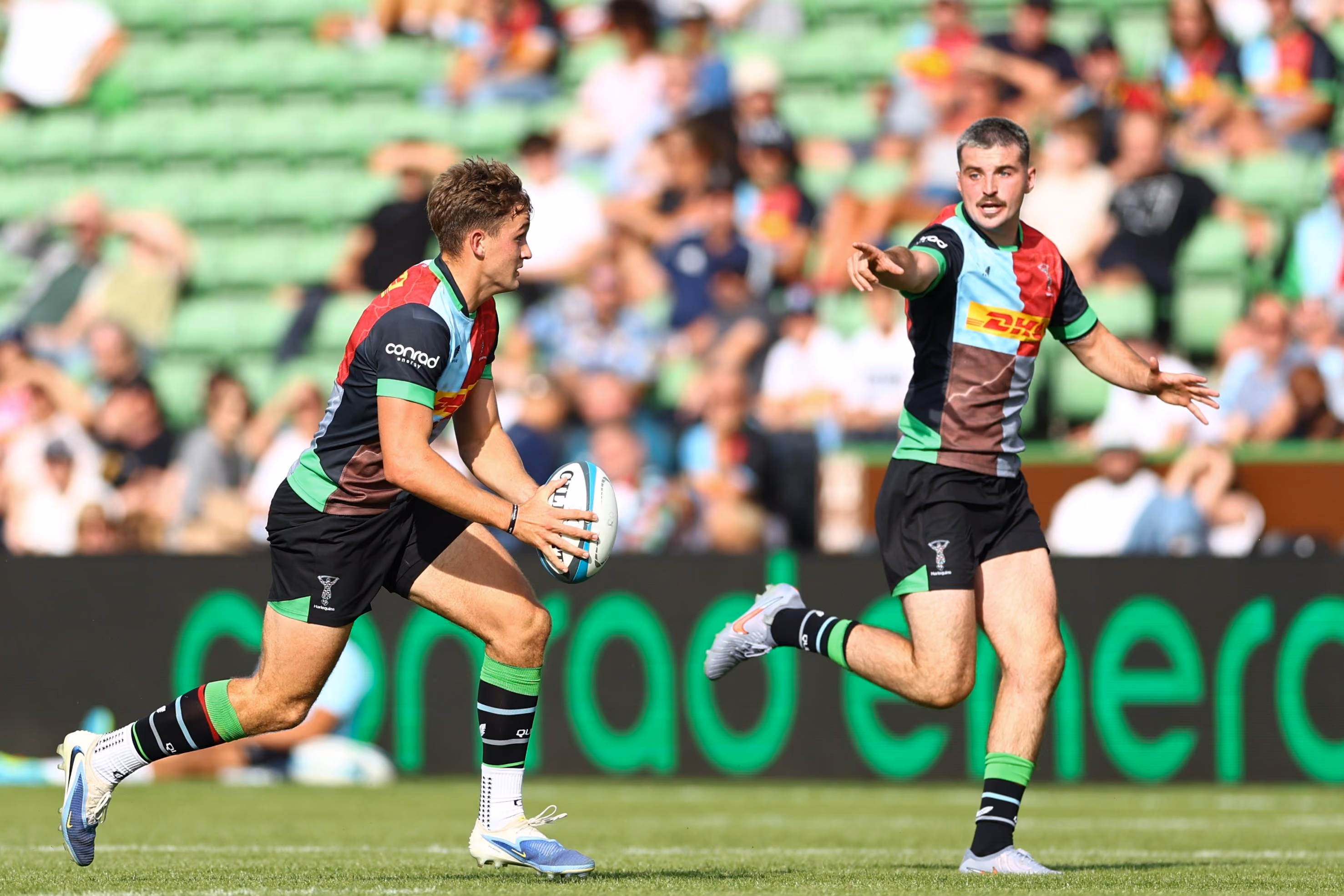 Two rugby players in Harlequins uniforms passing the ball during a match on a sunny day.