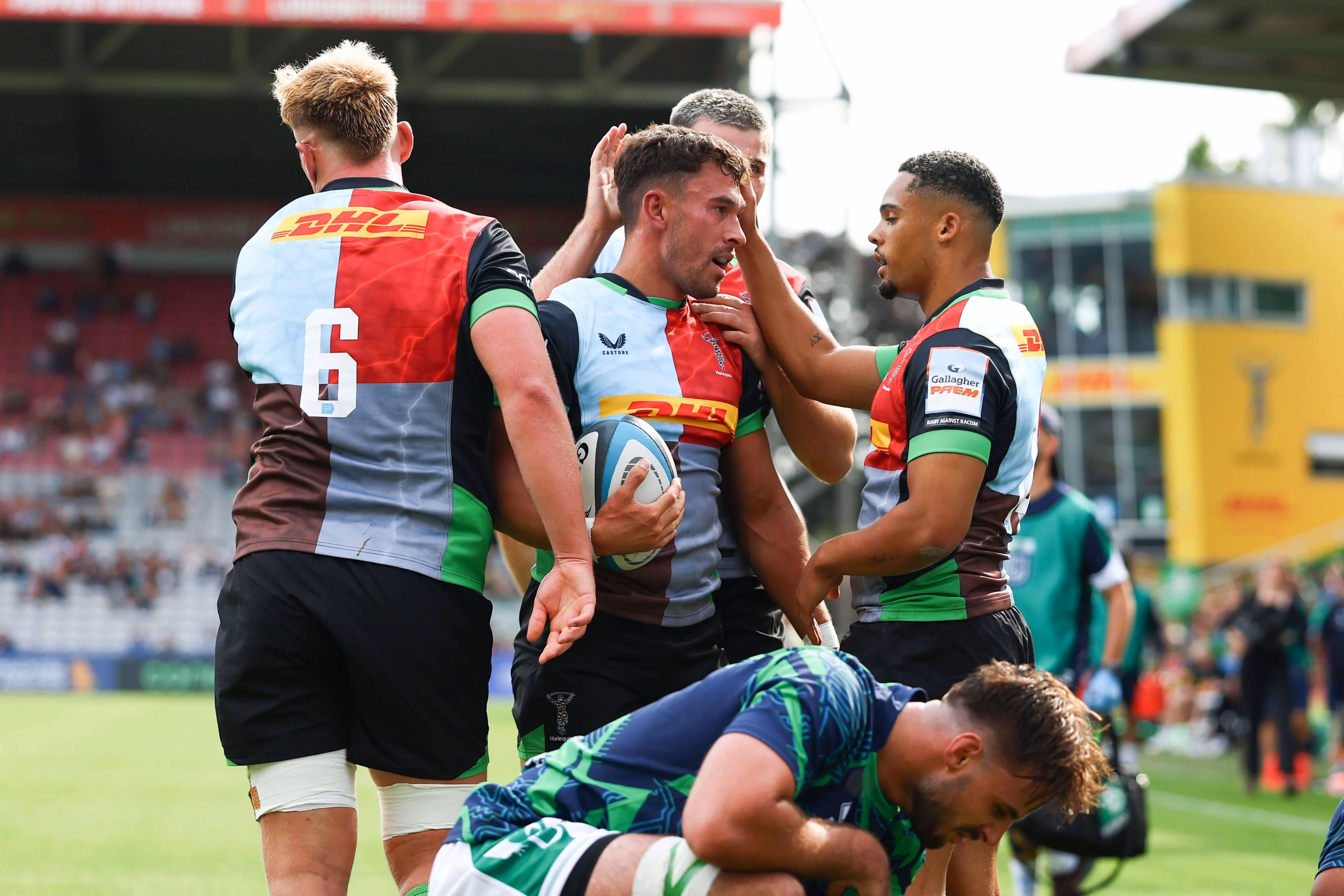 Four rugby players from a team in multicolored jerseys celebrating on the field with one holding a rugby ball, while a player in blue and green lies on the ground.