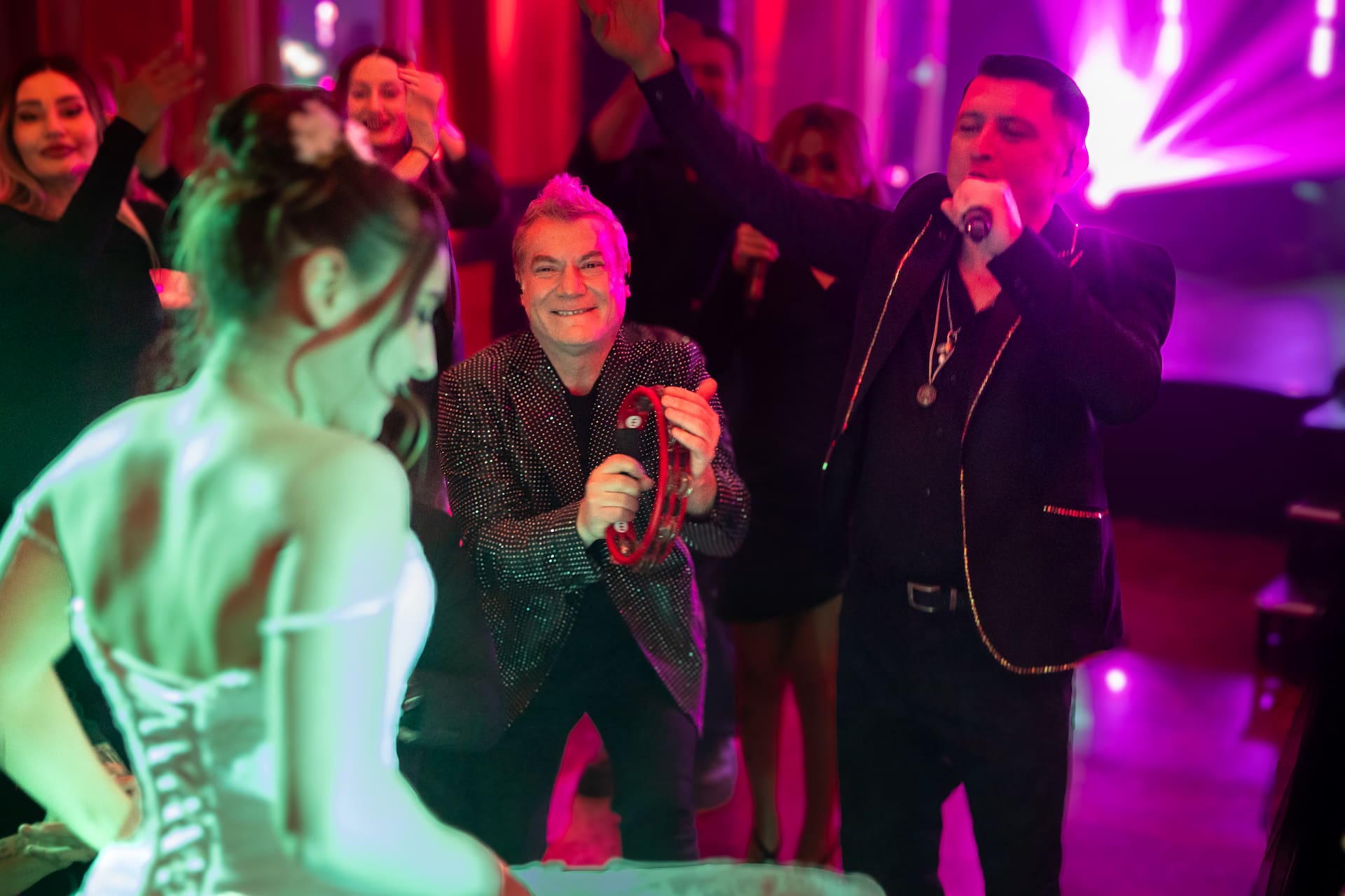 Bărbat cântând la microfon și altul dansând cu tamburină la o petrecere cu lumină roz și oameni care se distrează.