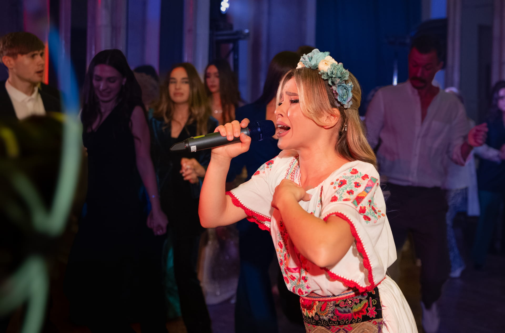 Femeie cântând la microfon într-o cămașă tradițională brodată, cu coroană de flori pe cap, în fața unui grup de oameni la un eveniment.