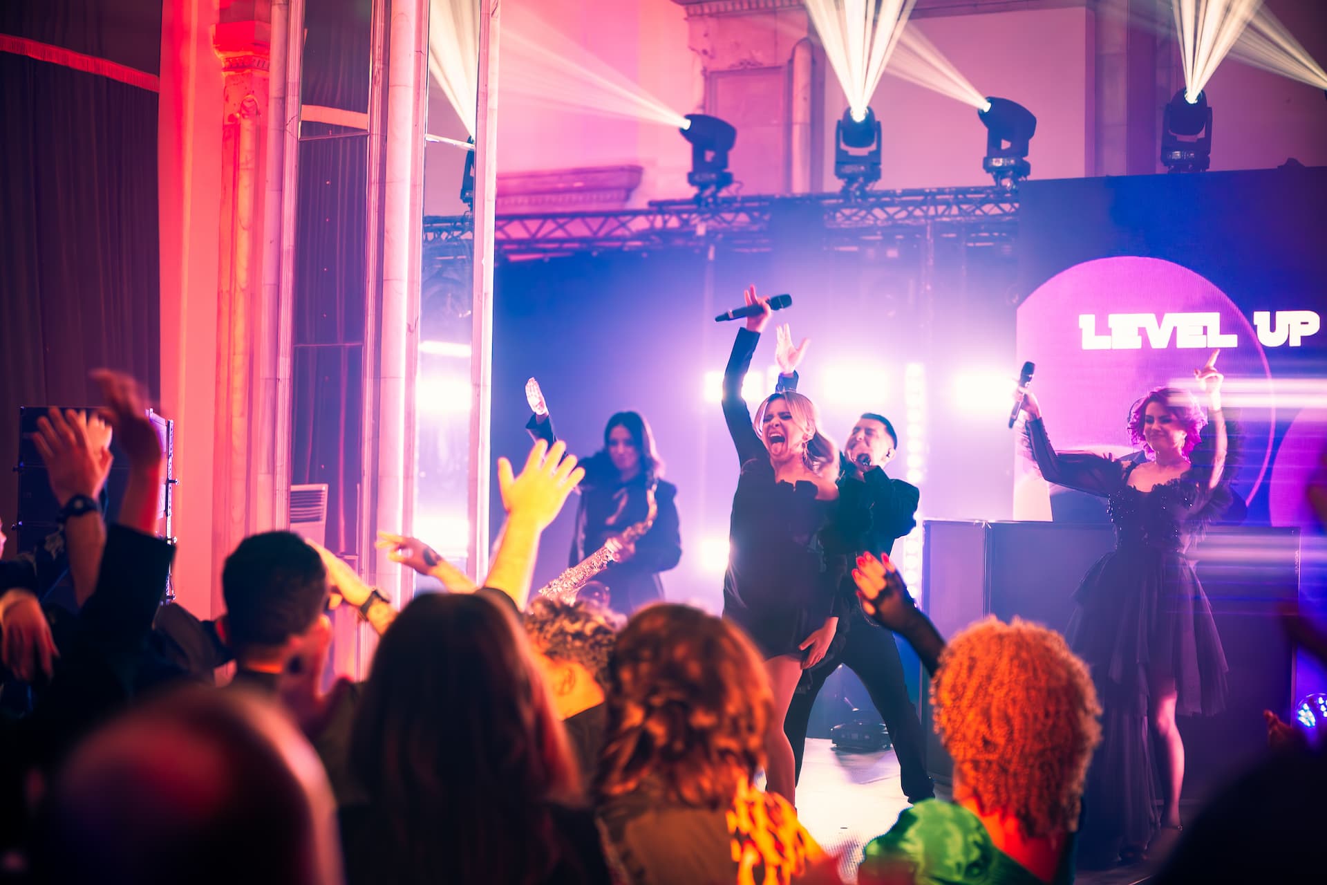 Artiști cântând și dansând pe scenă, cu publicul ridicând mâinile în sus, sub luminile colorate ale concertului.