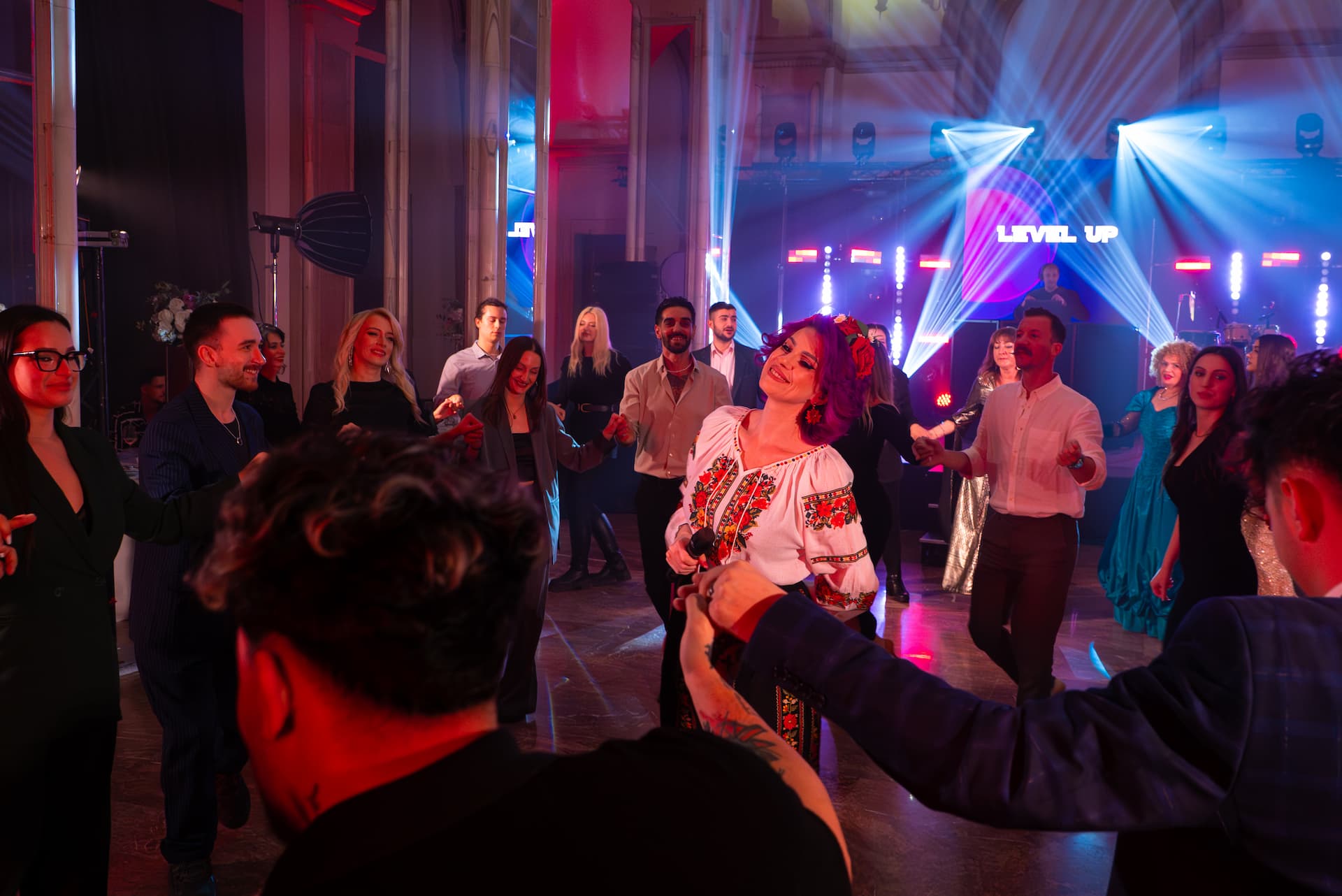 Un grup de oameni dansează în cerc într-un club cu lumină colorată și un ecran pe fundal cu textul "LEVEL UP".