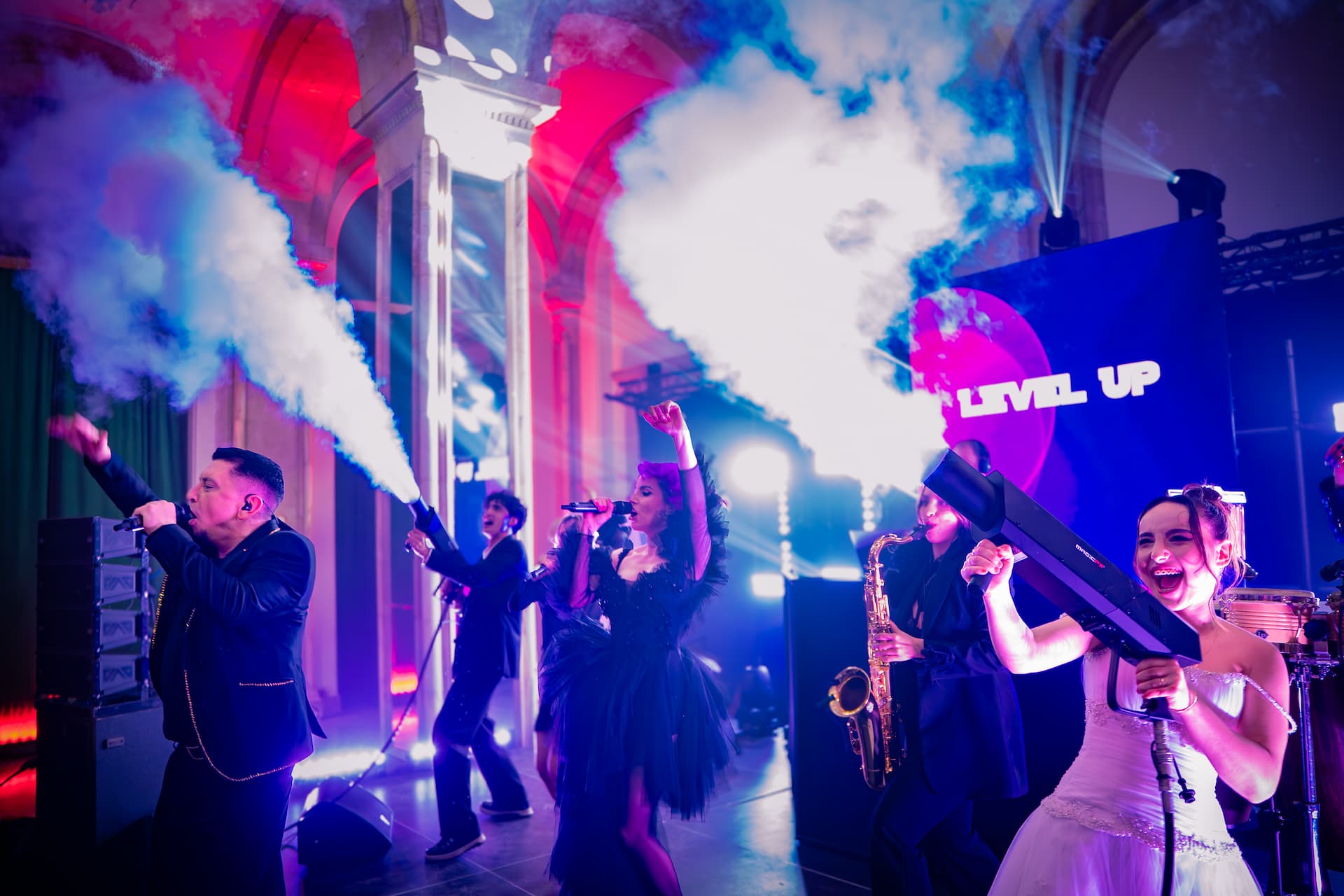 Artiști pe scenă la un concert vibrant cu efecte de fum și lumini colorate, inclusiv o femeie în rochie albă ținând un tun de fum.