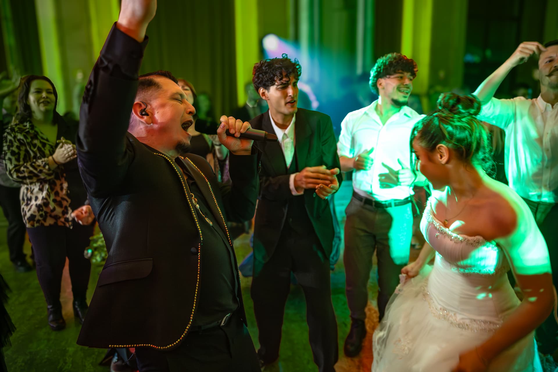 Oameni la o petrecere, un bărbat cântă la microfon și o mireasă dansează cu invitații în jur, lumini verzi în fundal.