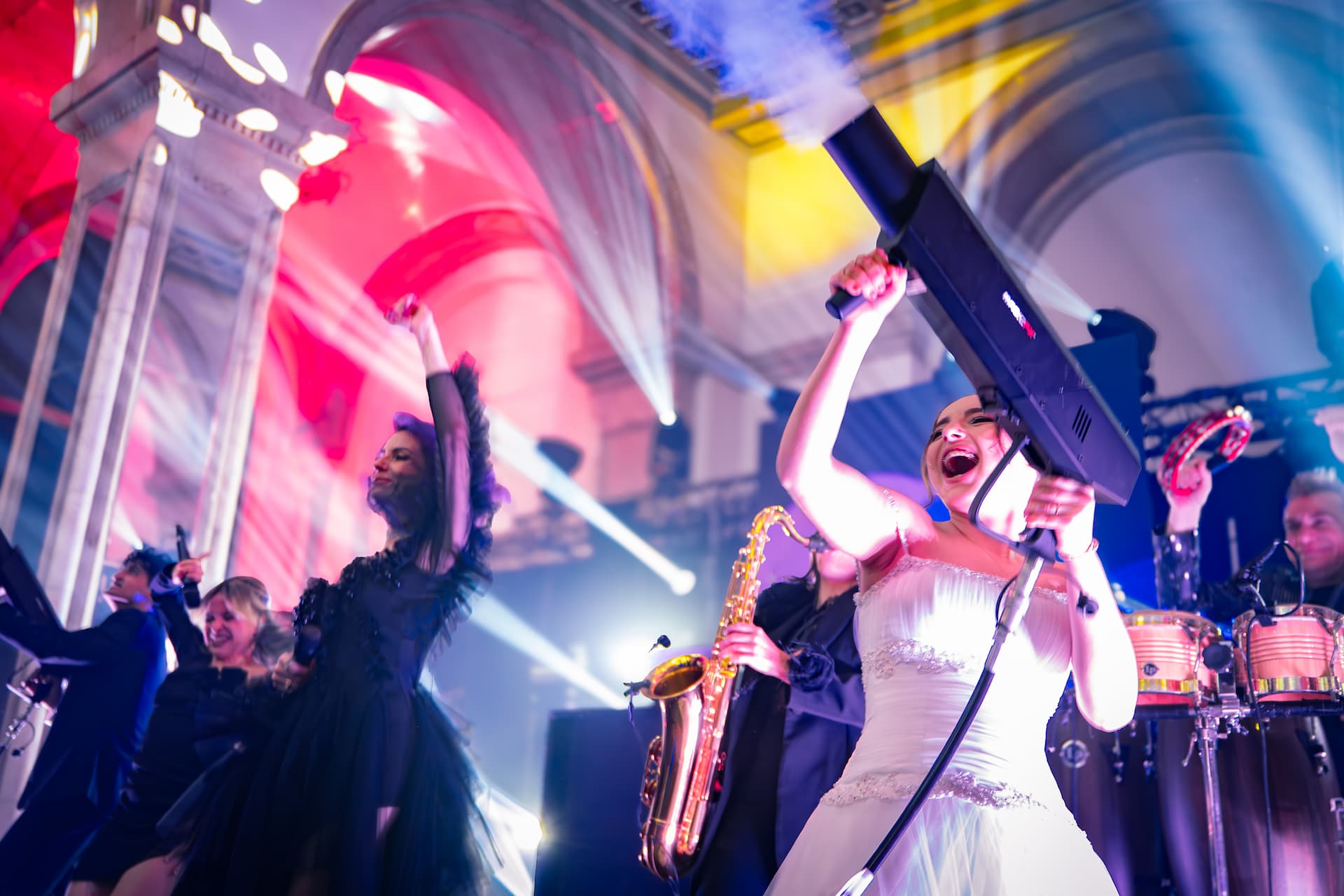 Femeie într-o rochie albă de mireasă ținând un dispozitiv de ceață pe o scenă colorată, înconjurată de muzicieni care cântă la saxofon și tobe, într-o atmosferă festivă cu lumini neon.