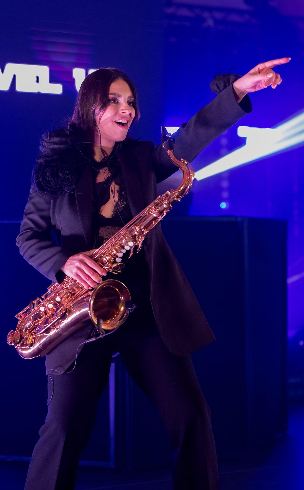 Femeie cântând la saxofon pe scenă, îmbrăcată elegant, în timp ce arată entuziastă și indică cu mâna dreaptă.