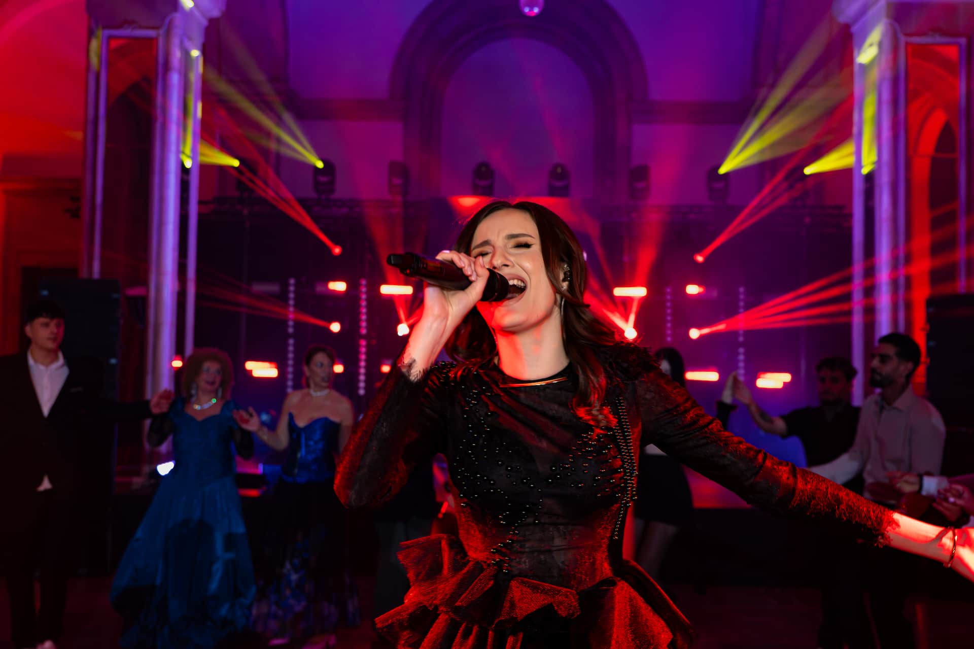 Femeie cântând cu microfon într-o rochie neagră elegantă, luminată de lumini roșii, cu oameni dansând în fundal.