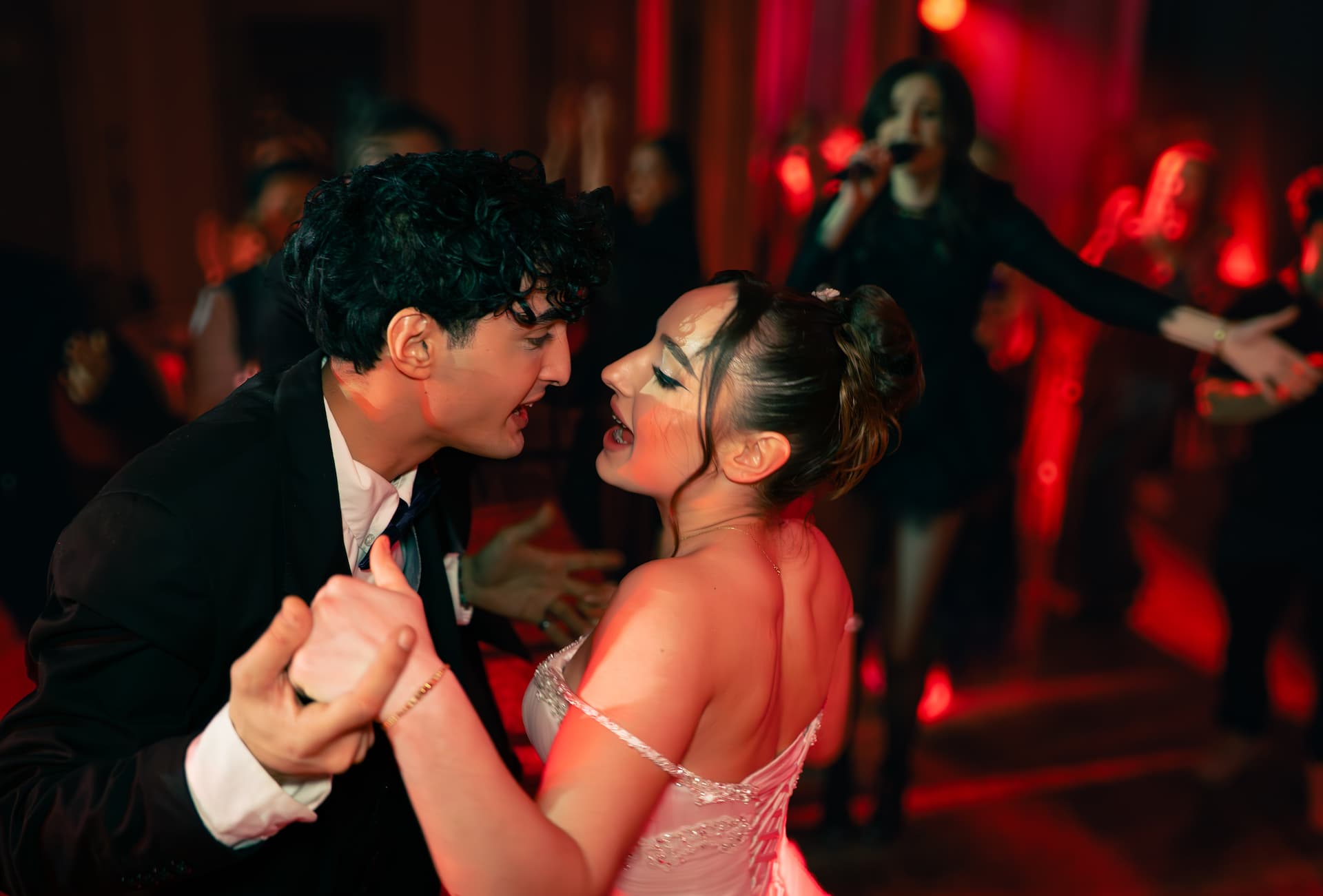 Un cuplu dansează îmbrăcat elegant, înconjurat de oameni care cântă și dansează într-un club cu lumină roșie.