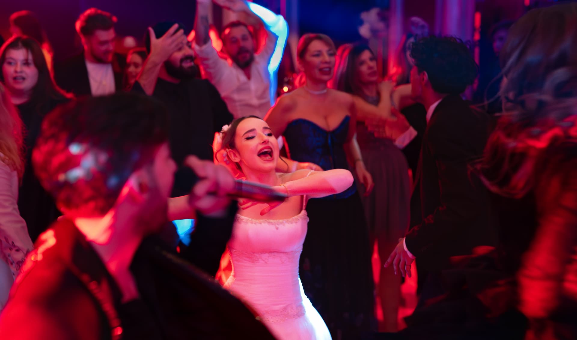 Mireasă veselă care cântă și dansează înconjurată de invitați la o petrecere cu lumină roșie vibrantă.