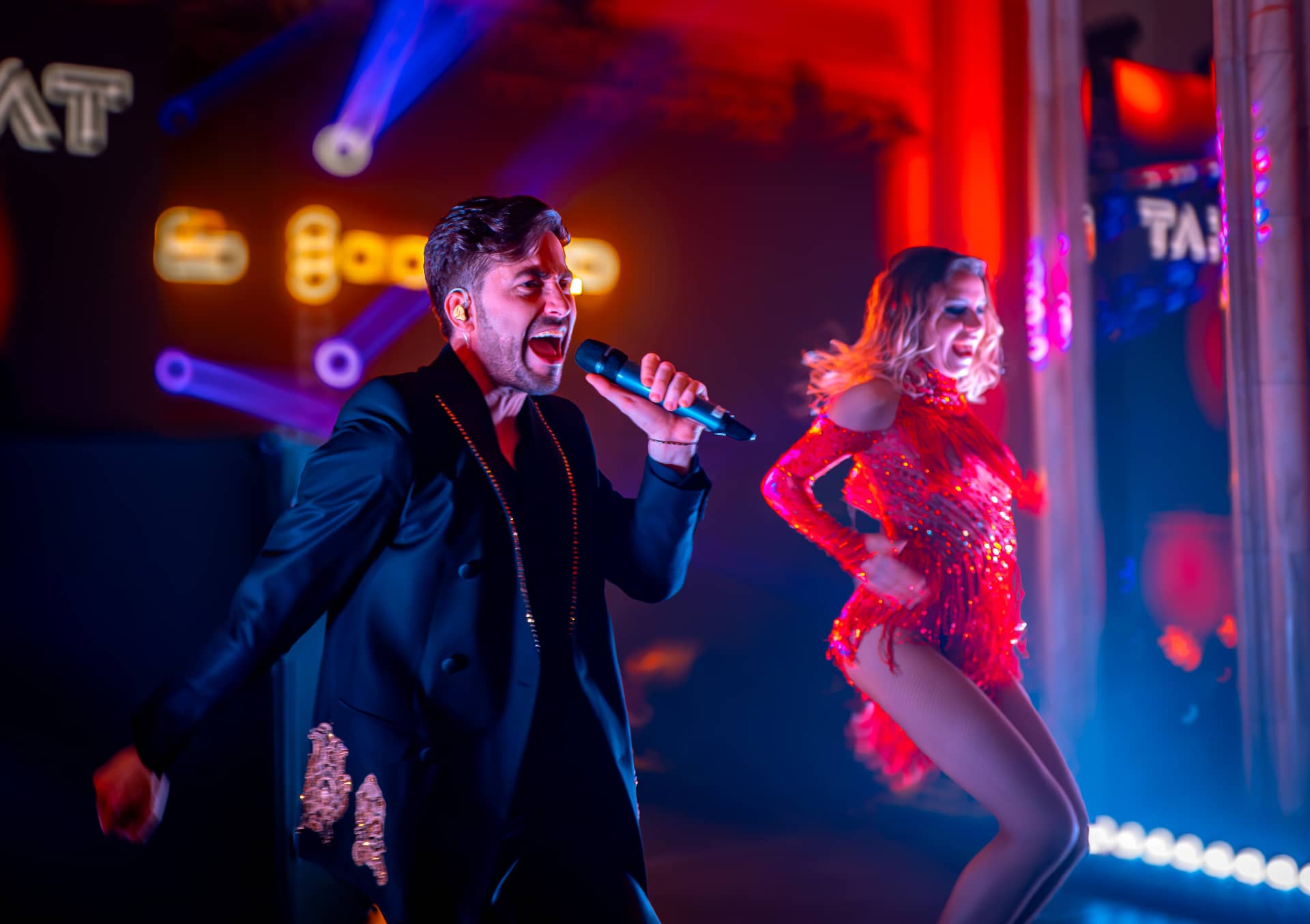 Bărbat cântând într-un microfon și femeie dansând într-un costum roșu sclipitor într-o scenă iluminată colorat.