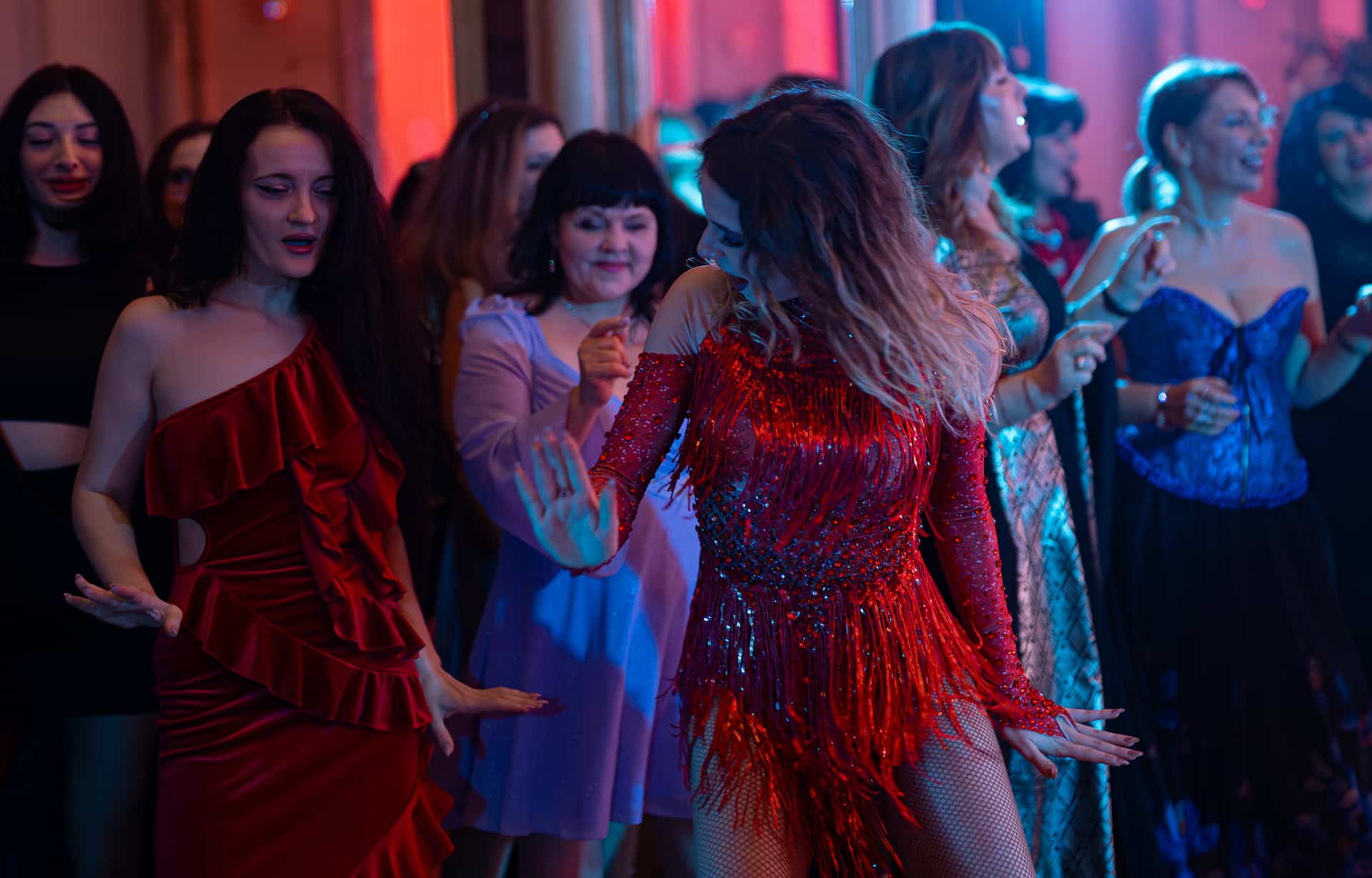 Femei dansând și distrându-se la o petrecere în lumini colorate, majoritatea purtând rochii elegante.