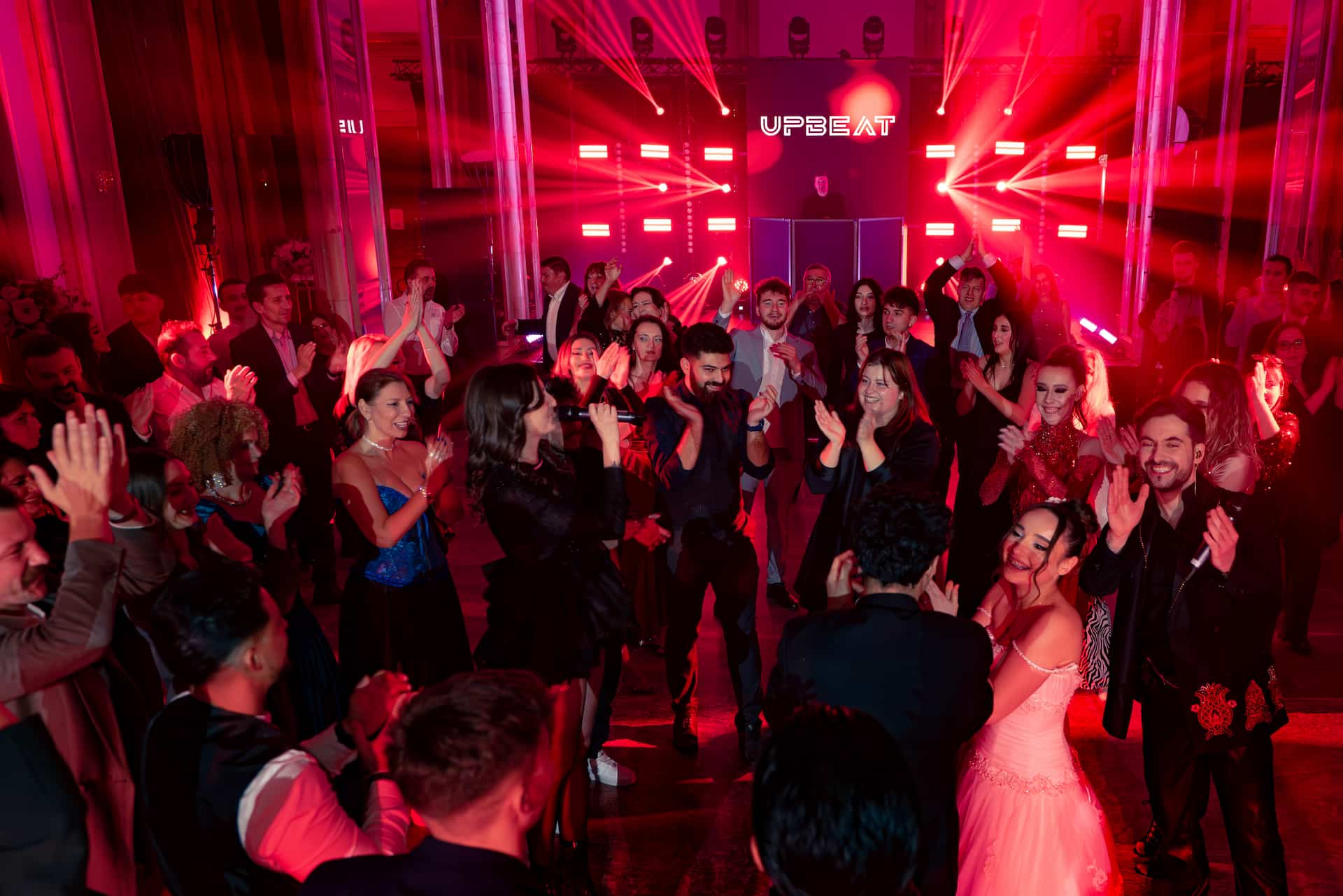 Oameni îmbrăcați elegant dansează și aplaudă într-un club cu iluminat roșu și DJ pe scenă.