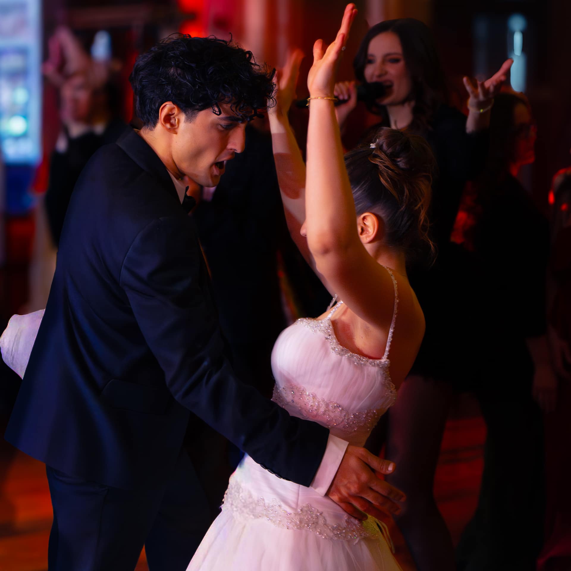 Un cuplu dansează într-un decor festiv, femeia poartă o rochie albă elegantă, iar bărbatul un costum negru.