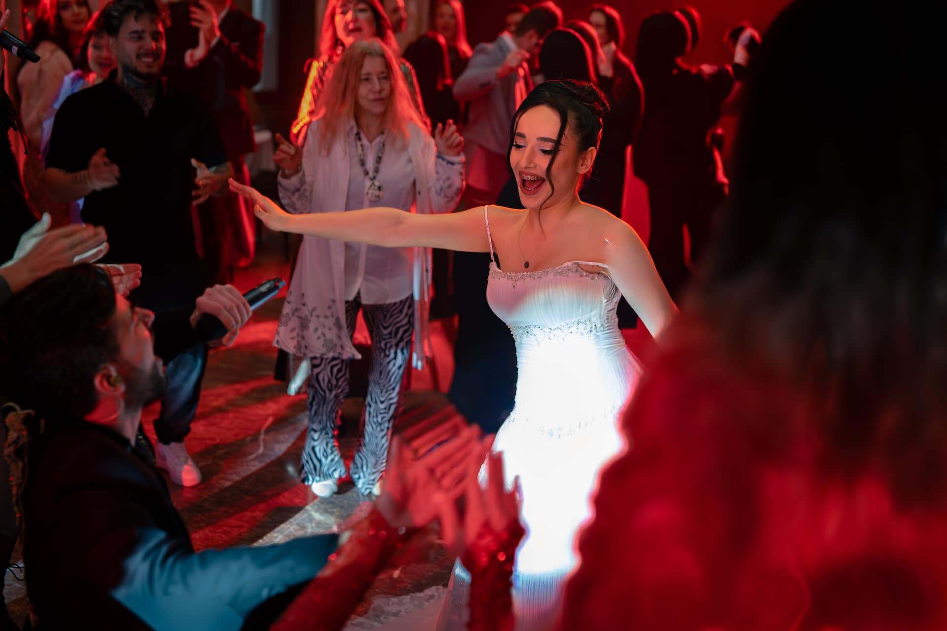 Femeie îmbrăcată într-o rochie albă dansează fericită înconjurată de o mulțime de oameni care aplaudă și se distrează într-un spațiu cu iluminare roșie.