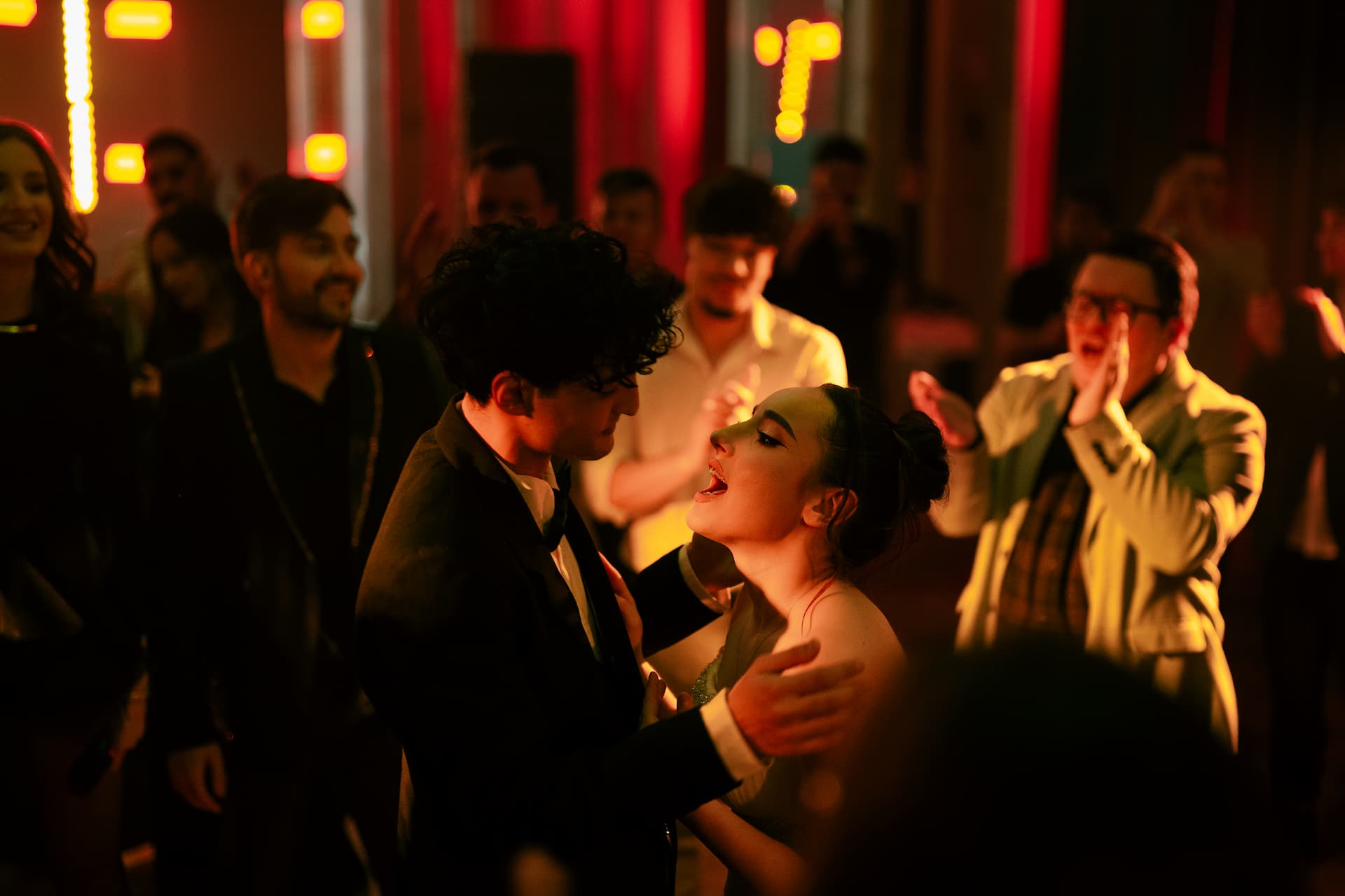 Un cuplu tânăr dansează și cântă împreună într-un club aglomerat, înconjurați de oameni care aplaudă și se distrează.