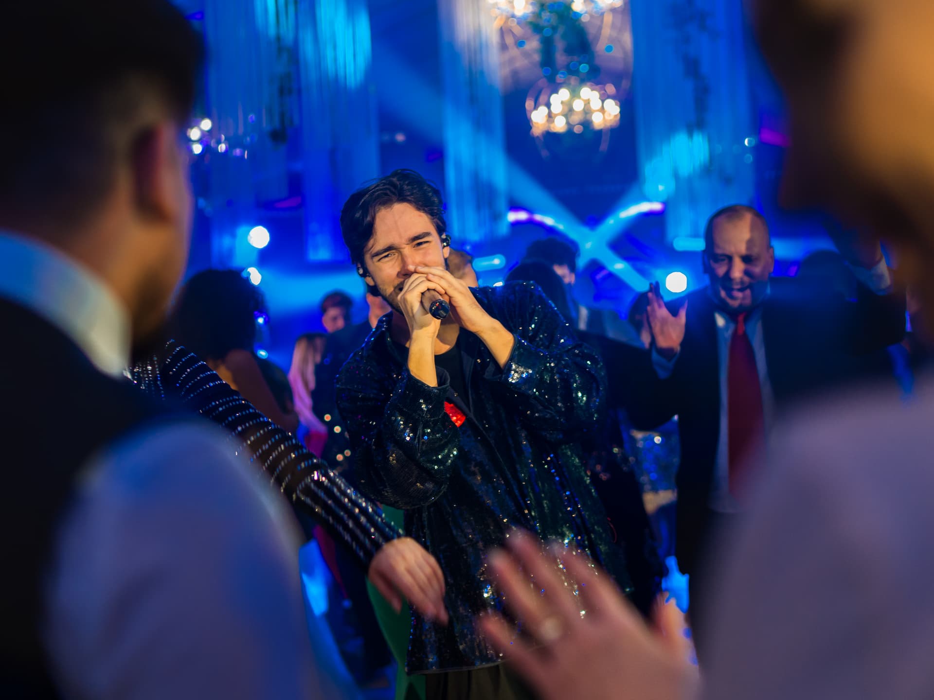 Un bărbat cântă la microfon într-un club de noapte cu lumini albastre, în timp ce mulțimea dansează și aplaudă în jur.