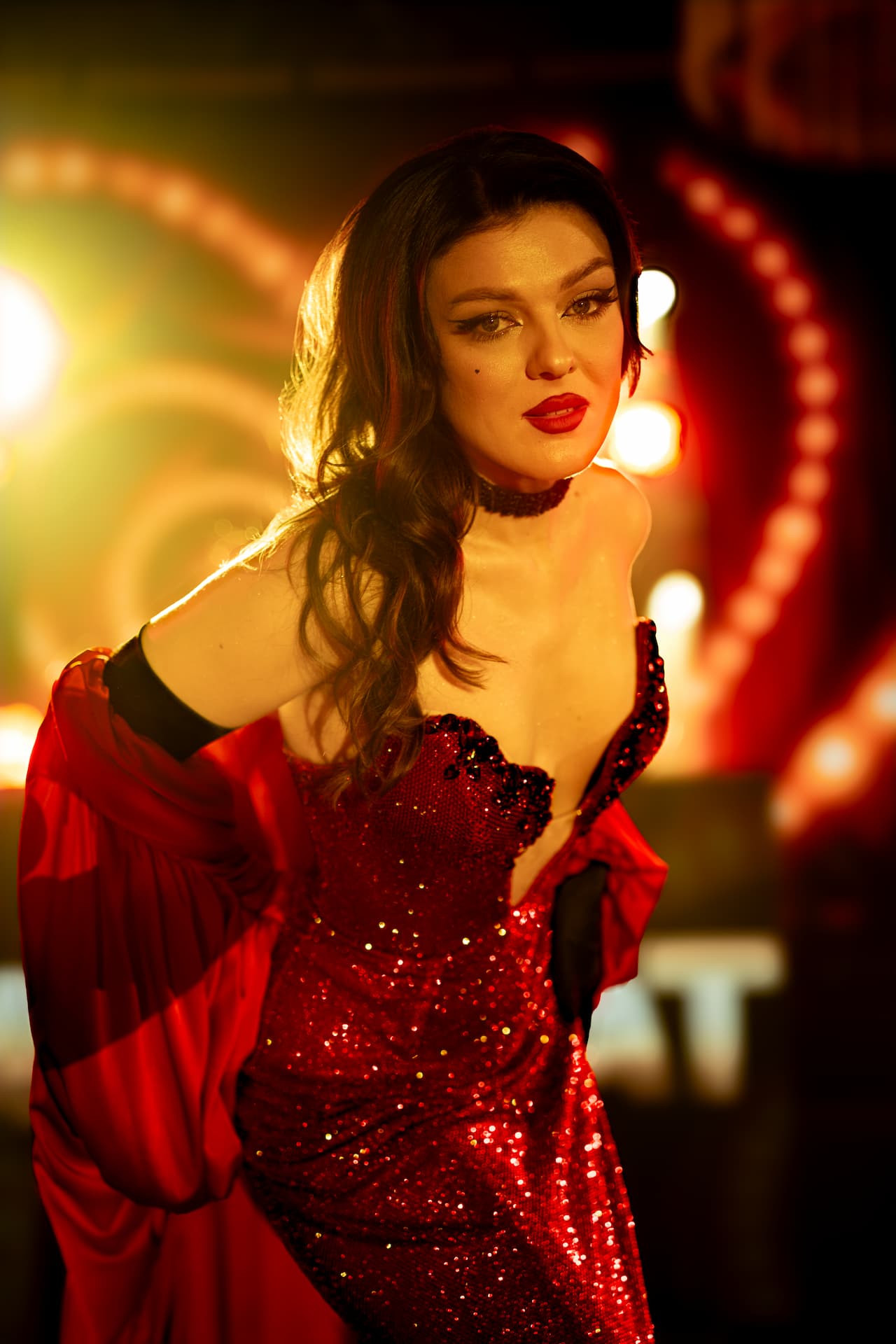 Femeie cu păr lung și machiaj dramatic poartă o rochie roșie sclipitoare și un șal roșu într-un decor cu lumini calde.