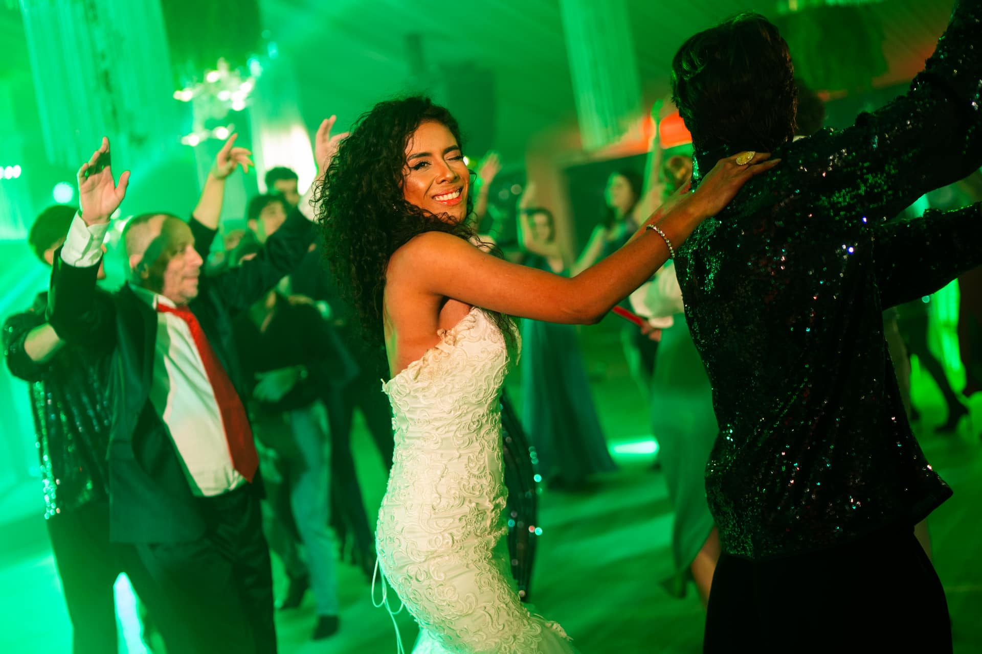 Fată îmbrăcată într-o rochie de mireasă albă dansează zâmbind la un eveniment cu mulți oameni în fundal, sub lumină verde.