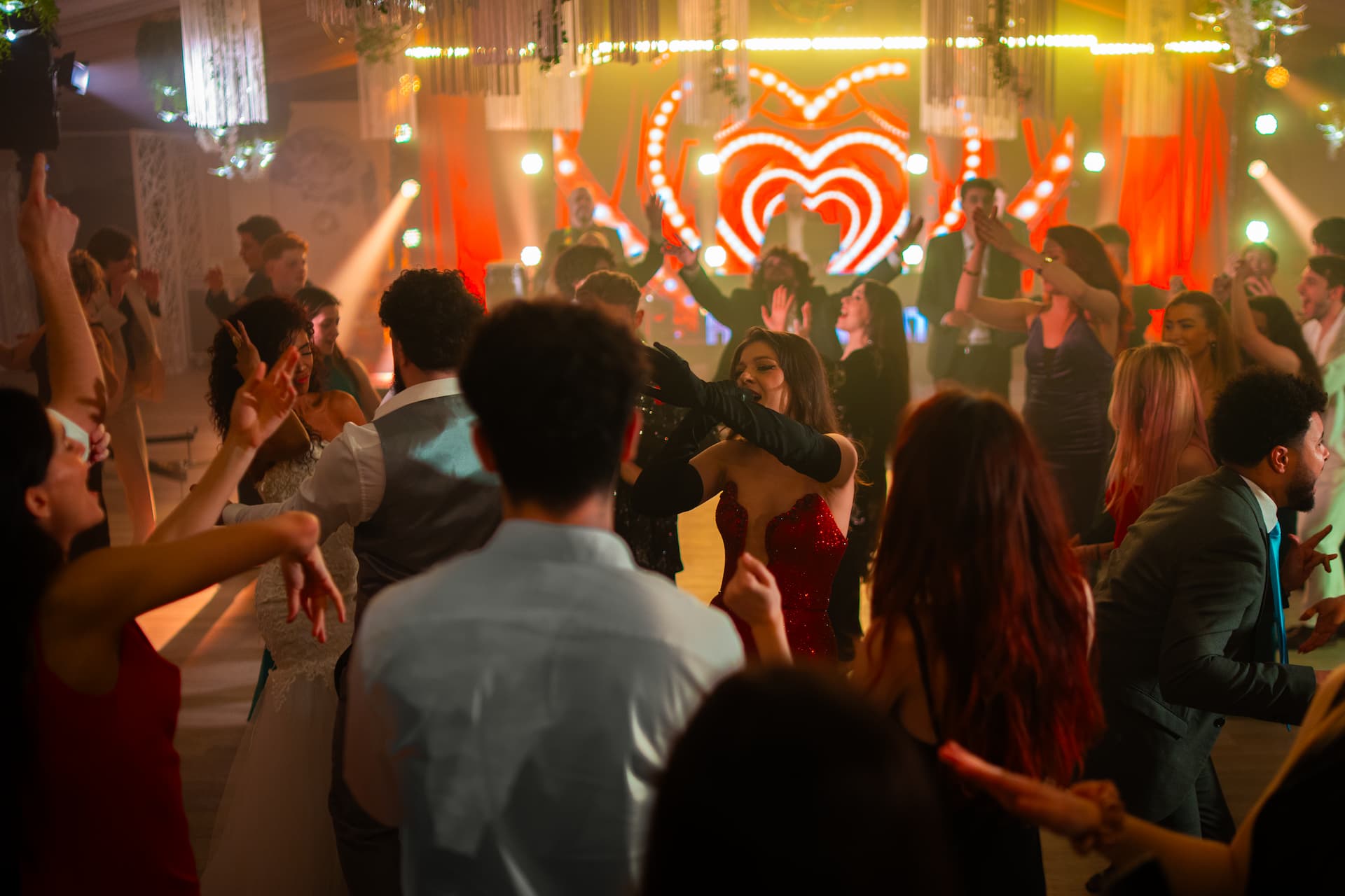 O mulțime de oameni dansează și se distrează într-un club cu lumină caldă și decorațiuni luminoase în formă de inimă pe fundal.